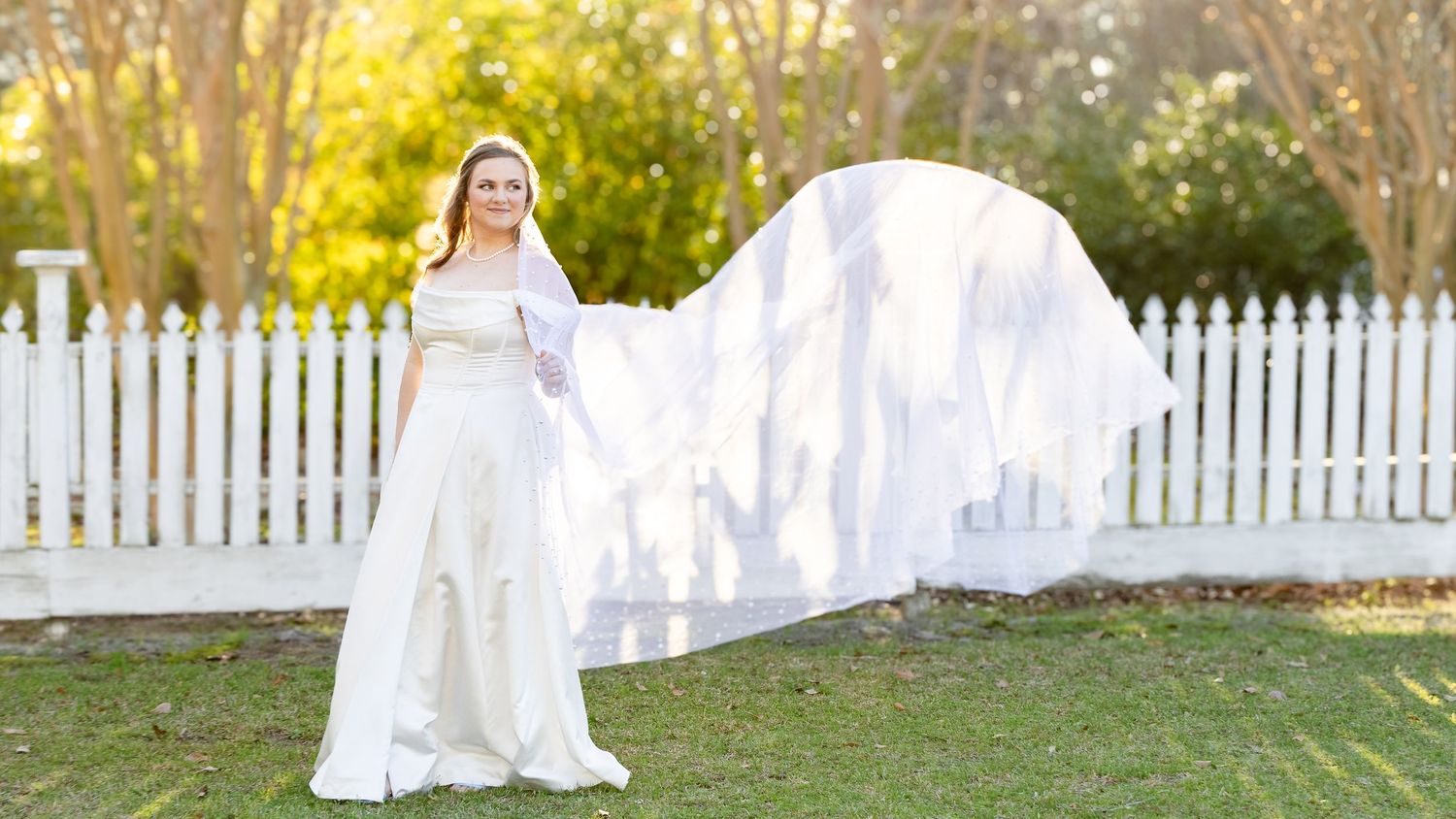 A white wedding dress hangs on a white picket fence in golden sunlight creating an ethereal outdoor display.