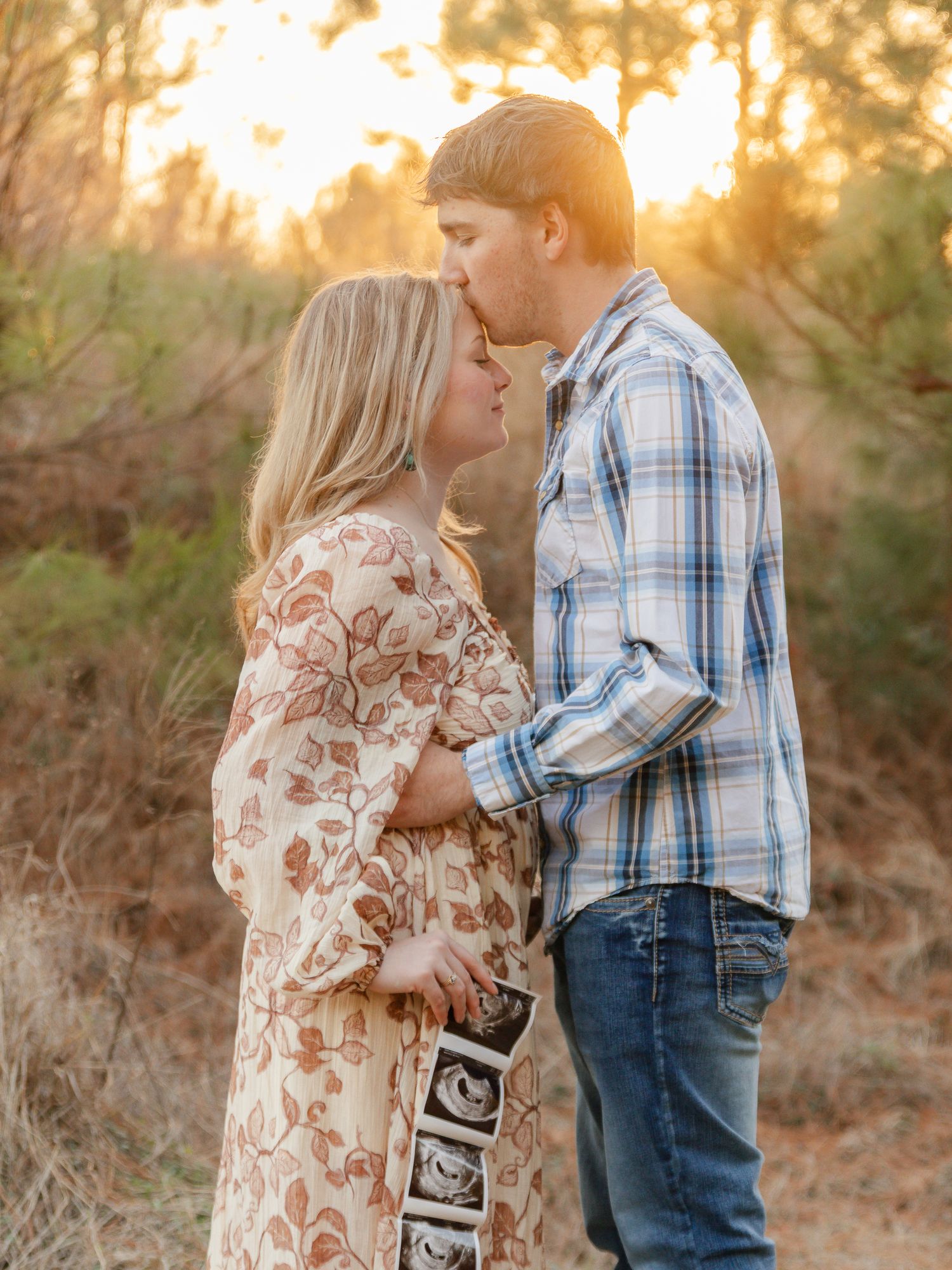 A couple shares an intimate moment at sunset while holding an ultrasound photo in a rustic outdoor setting.