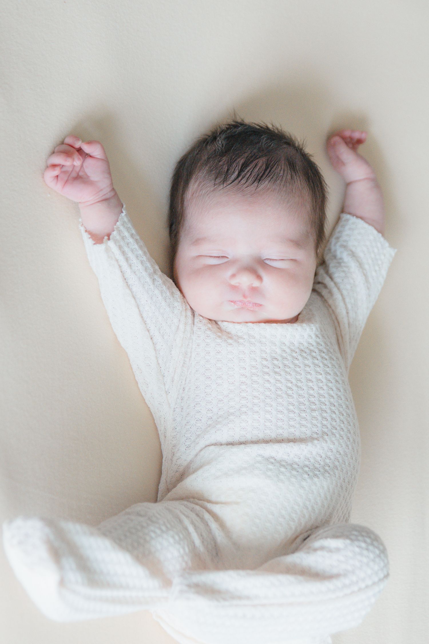 Newborn baby sleeping in white knit outfit with both arms raised up on cream background.