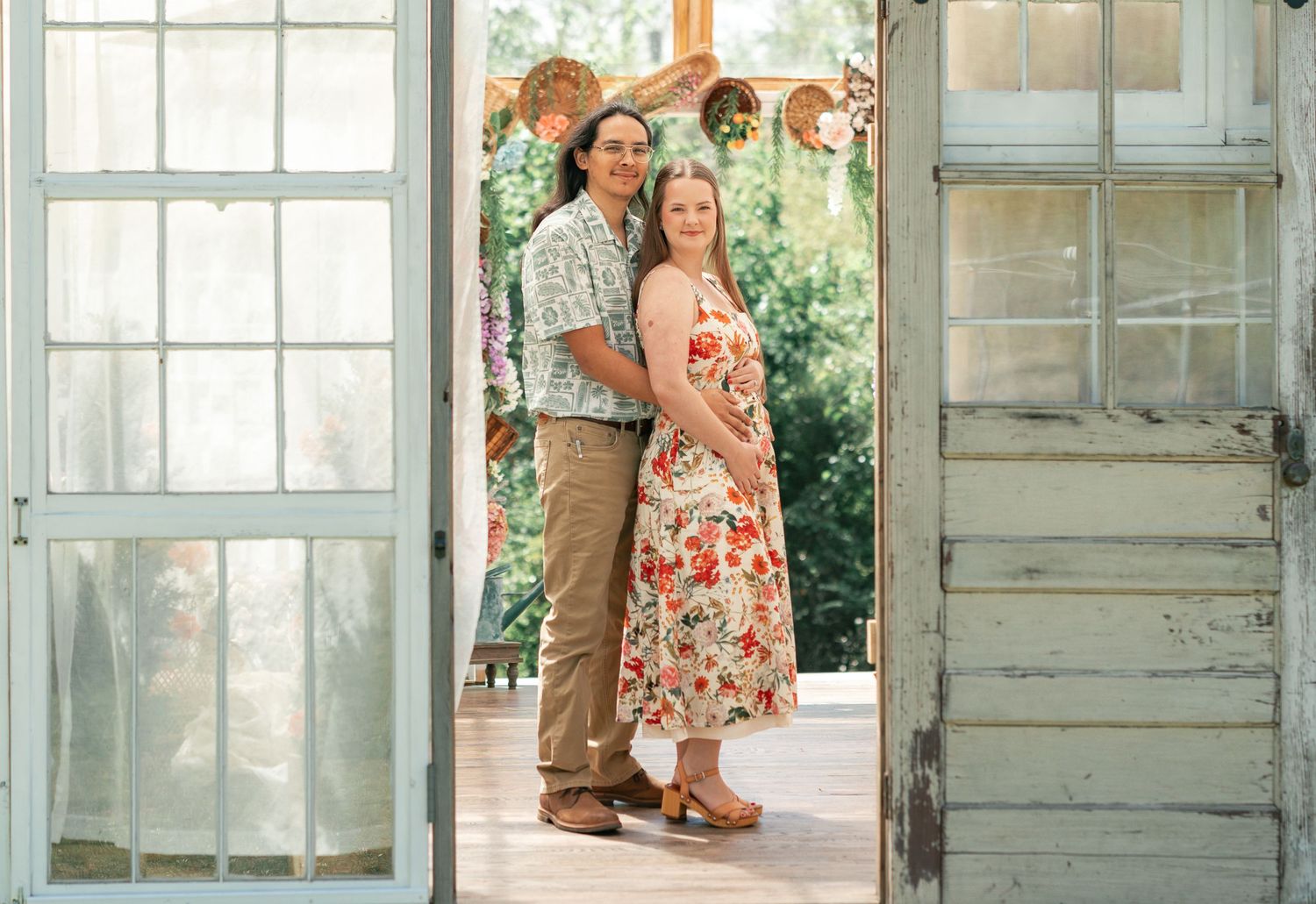 Couple posing together in a rustic doorway, woman in floral dress and man in casual attire, lush greenery background.