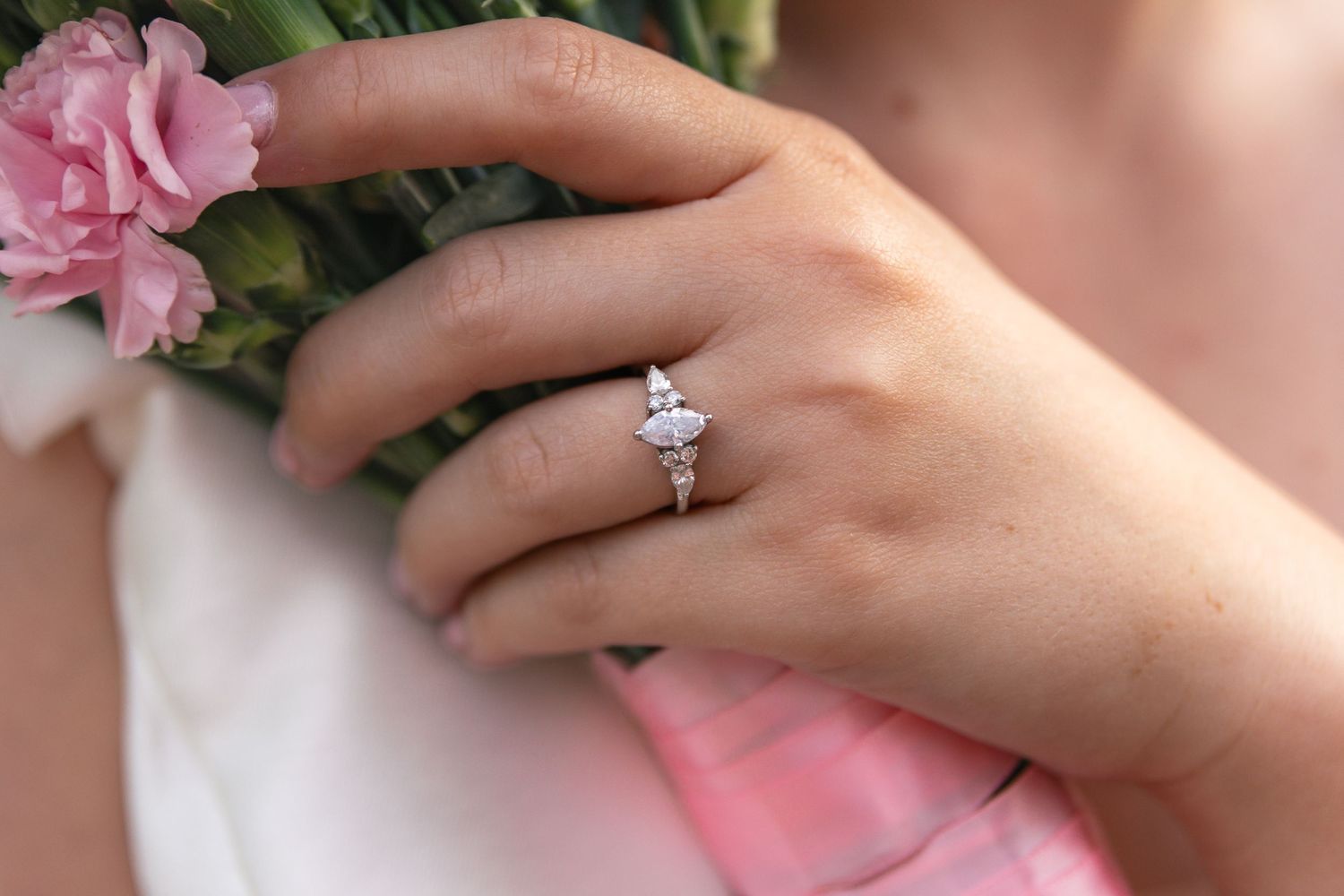 A sparkling diamond engagement ring shown on a hand next to pink roses and soft fabric.