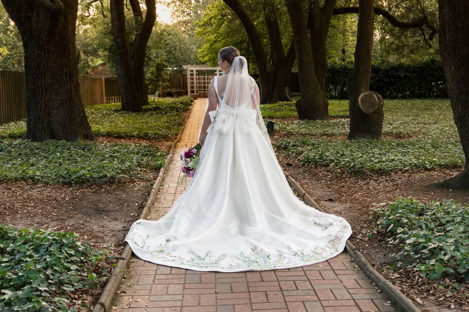A person in a flowing white wedding gown and veil stands in a wooded outdoor setting with dappled sunlight.