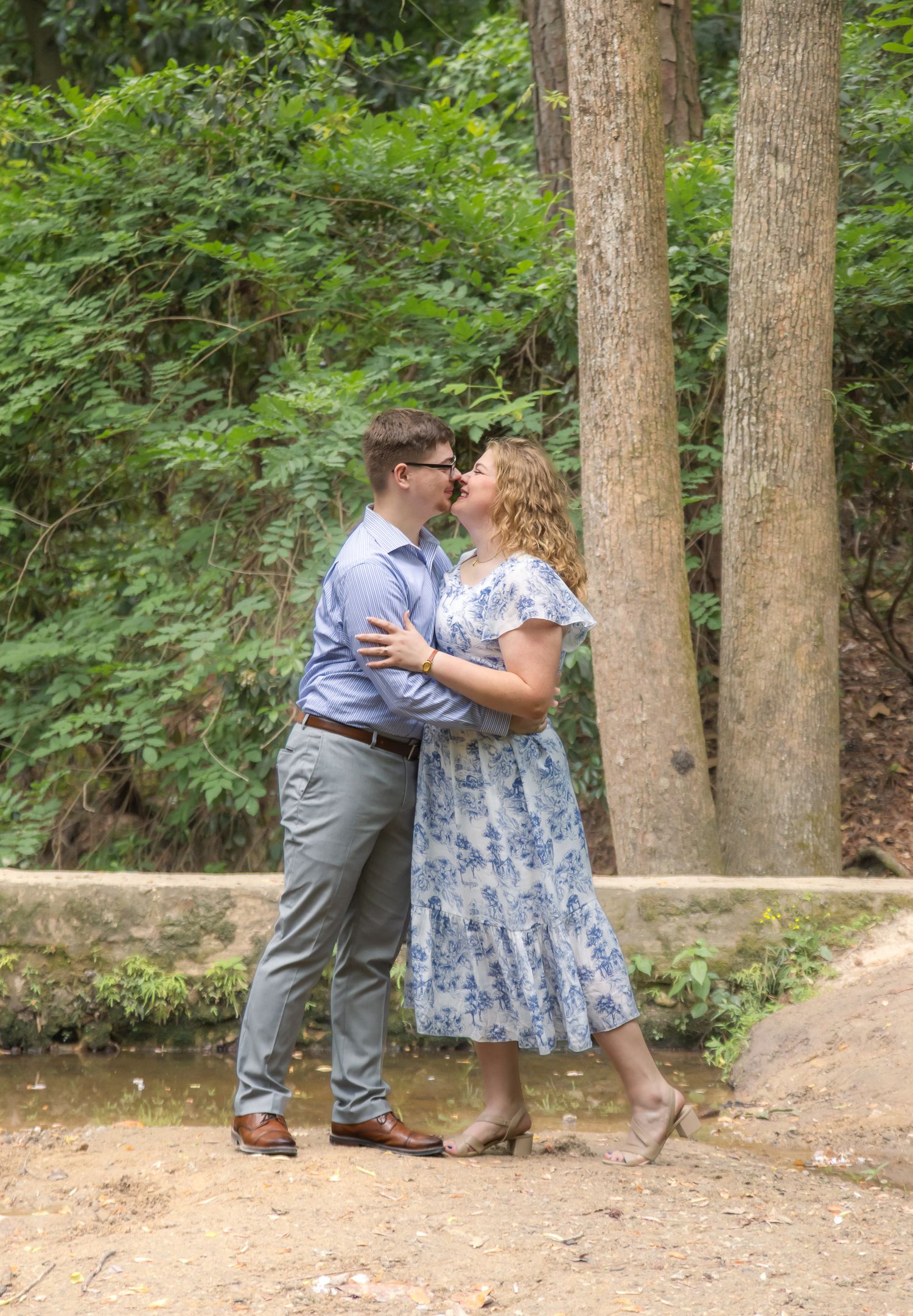 Couple sharing a kiss outdoors near a creek surrounded by lush green trees.