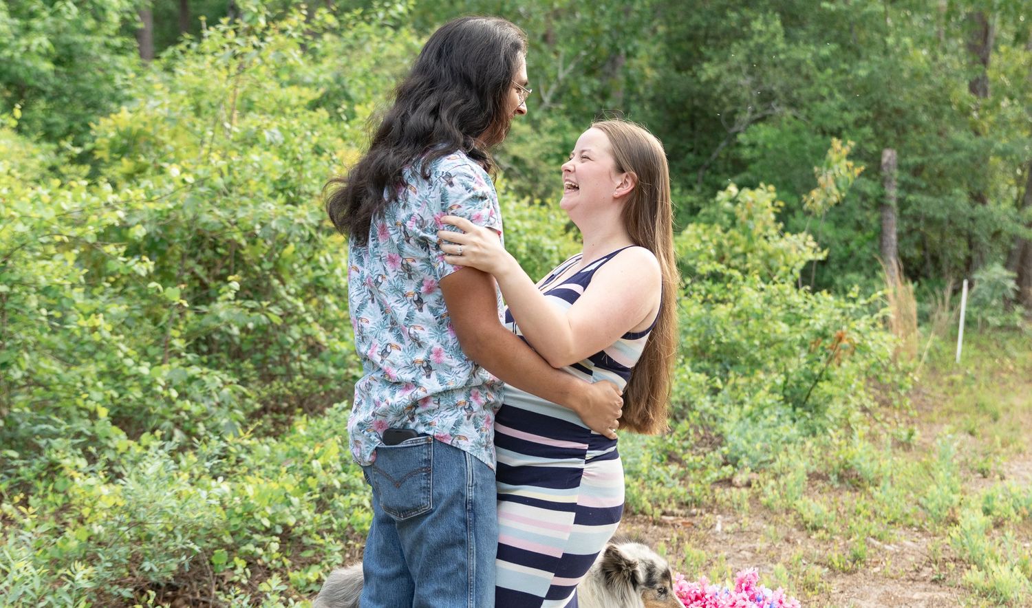 Couple embracing outdoors in a lush green setting, woman in striped dress appears pregnant, sharing a tender moment.