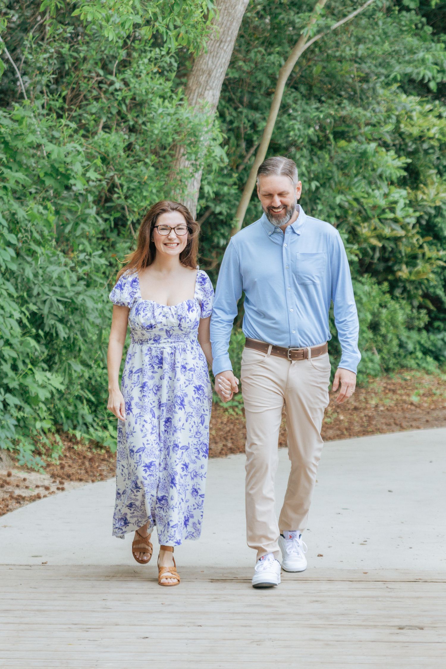 Couple holding hands walking on a path surrounded by lush green trees, woman in floral dress, man in blue shirt.