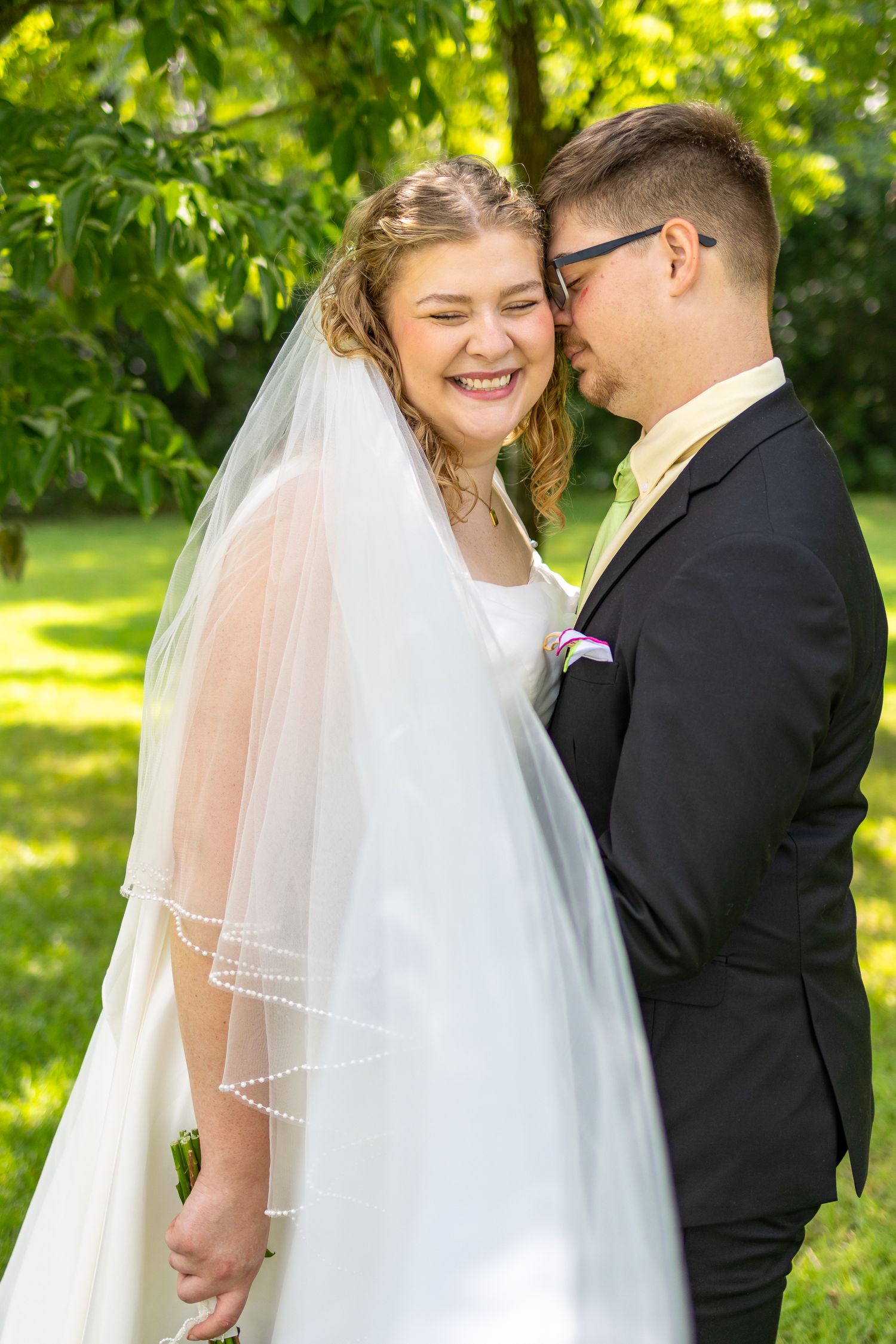 Happy bride in white veil laughs with groom in black suit outdoors in lush green garden setting.