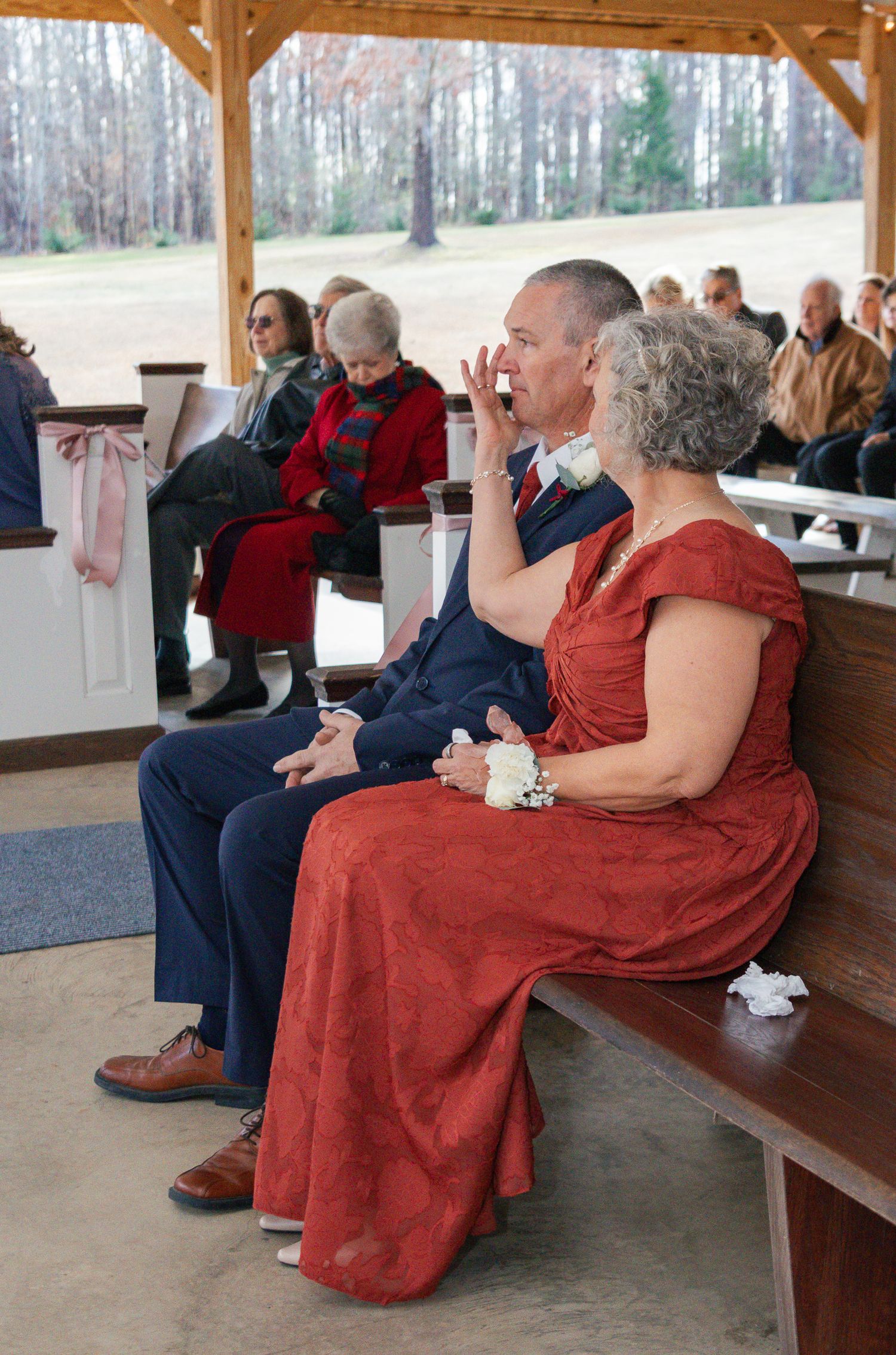 A group of seniors sitting on wooden benches at an outdoor covered pavilion watching an event together.
