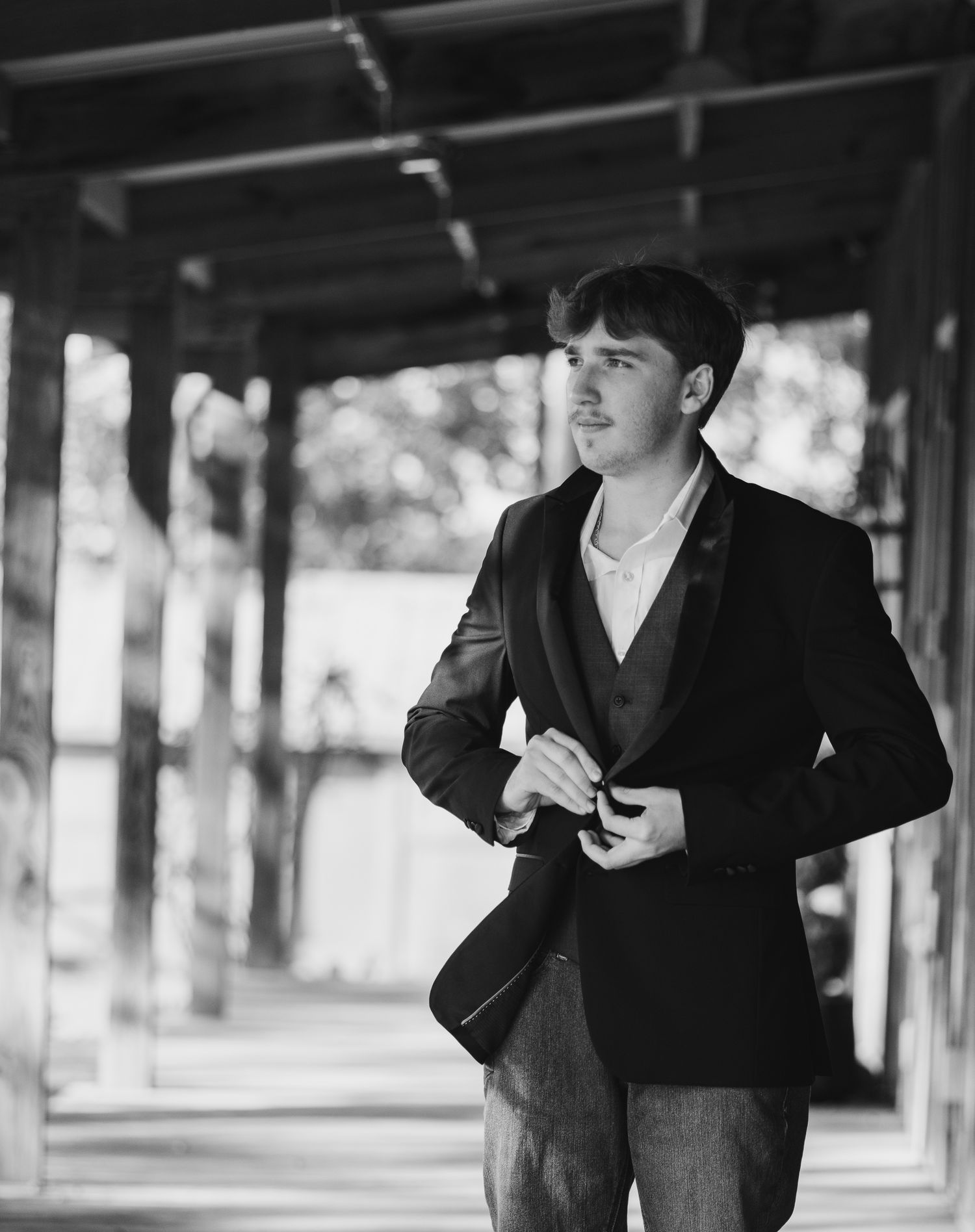 A person in a black suit stands in a covered walkway with pillars and trees visible in the background.
