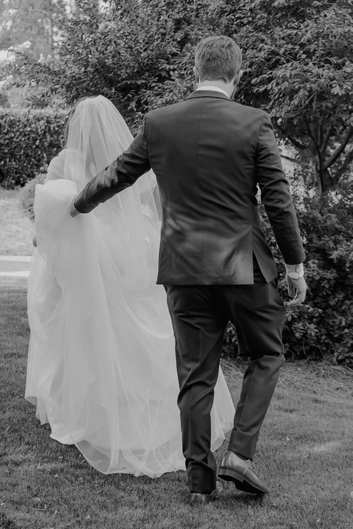 Black and white rear view of a bride in a veil and groom in a suit walking through a garden.