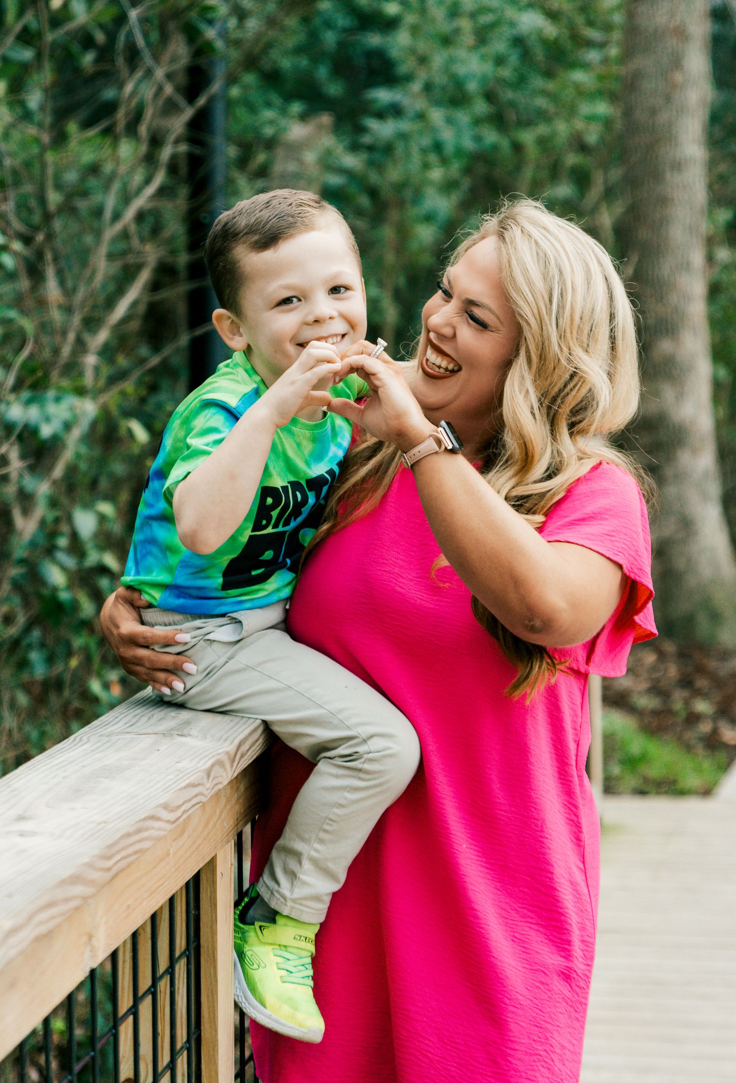 A laughing blonde woman in a pink dress holds a giggling young boy on a wooden deck outdoors.