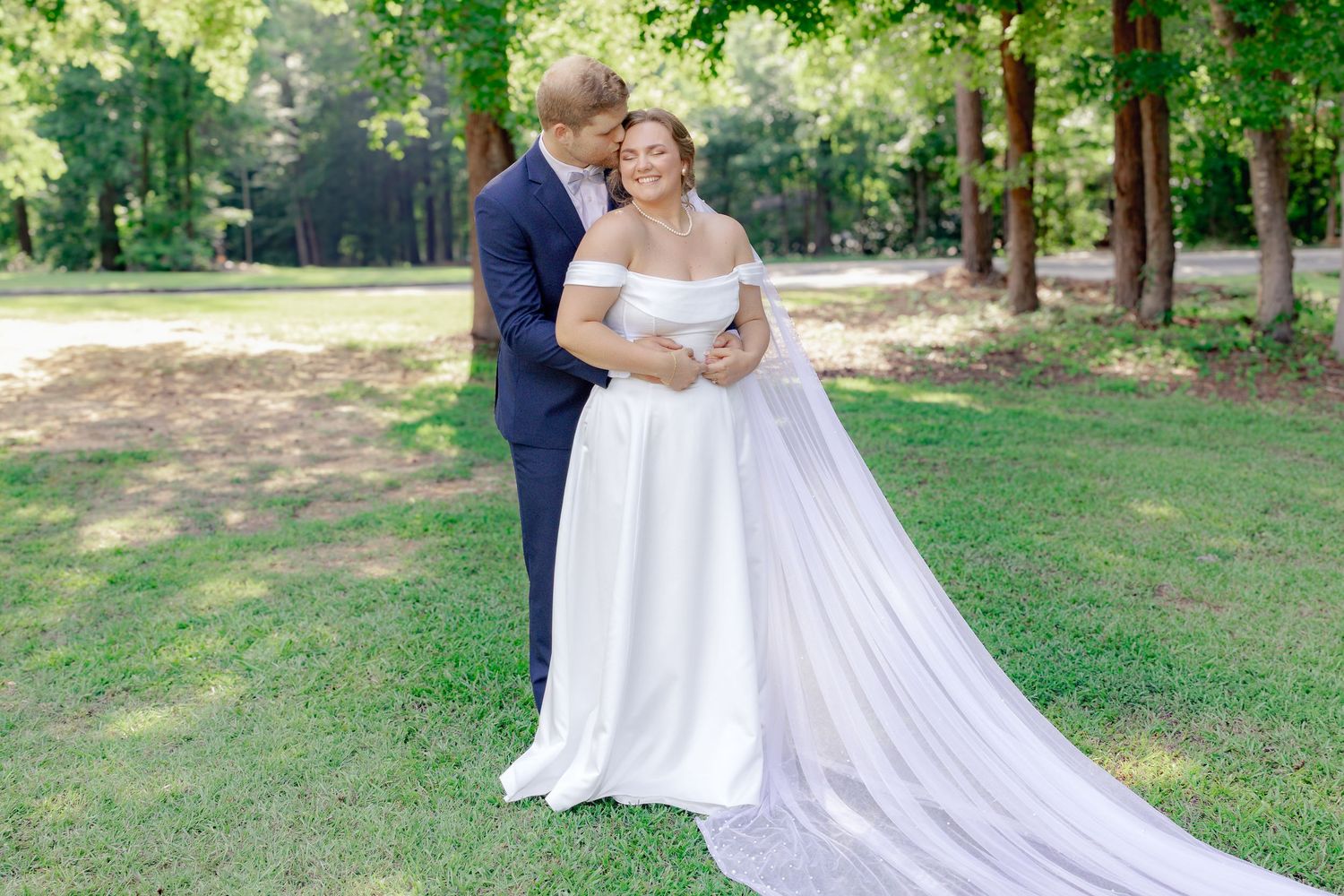 A couple in formal wedding attire poses together in a sunlit outdoor garden setting.