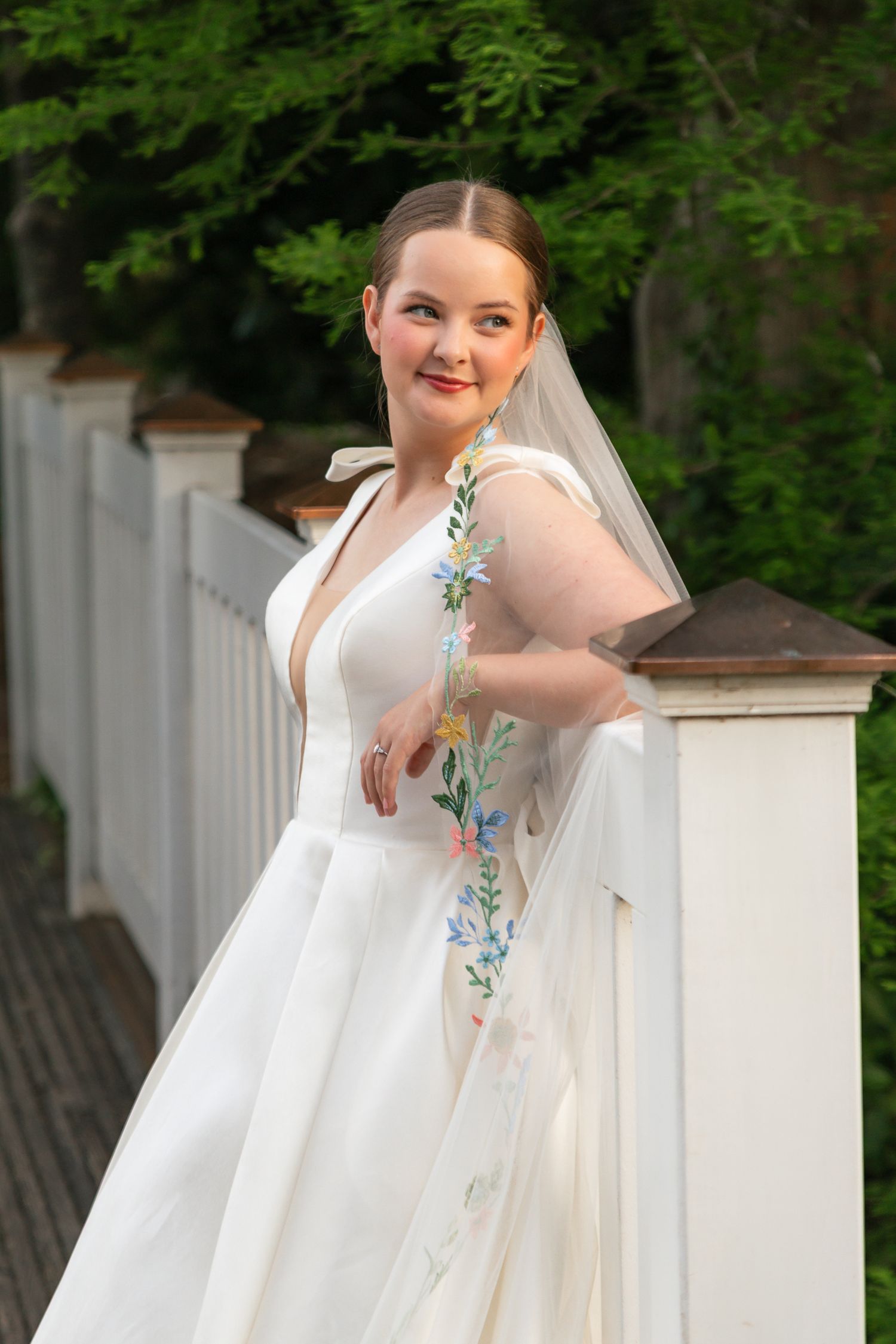 Bride in white gown with floral embroidered veil poses by white fence outdoors, smiling elegantly.