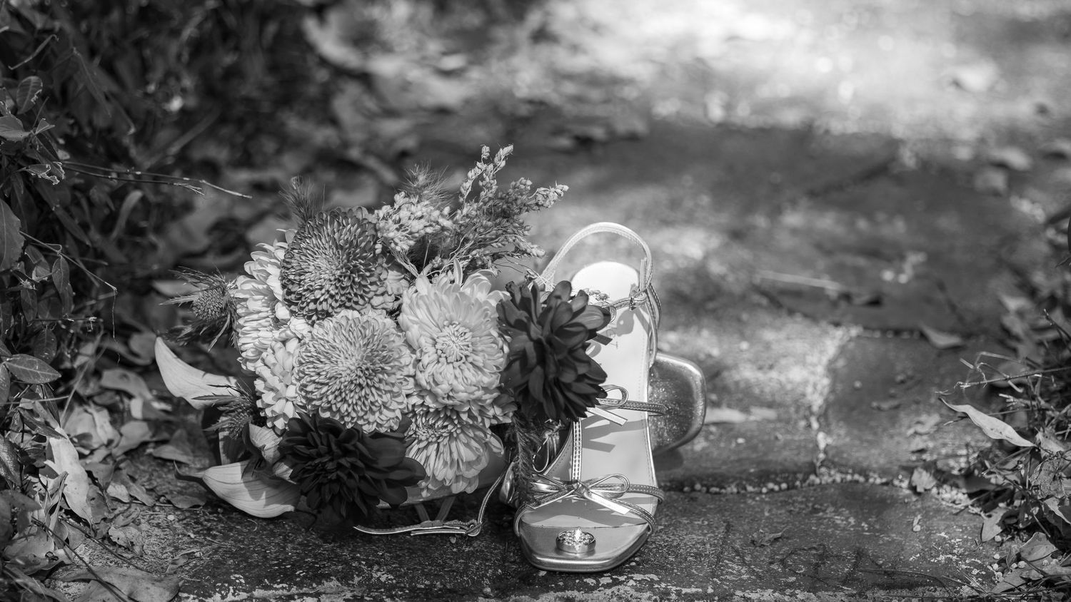 Black and white photo of a bridal bouquet resting beside a strappy heeled sandal outdoors.