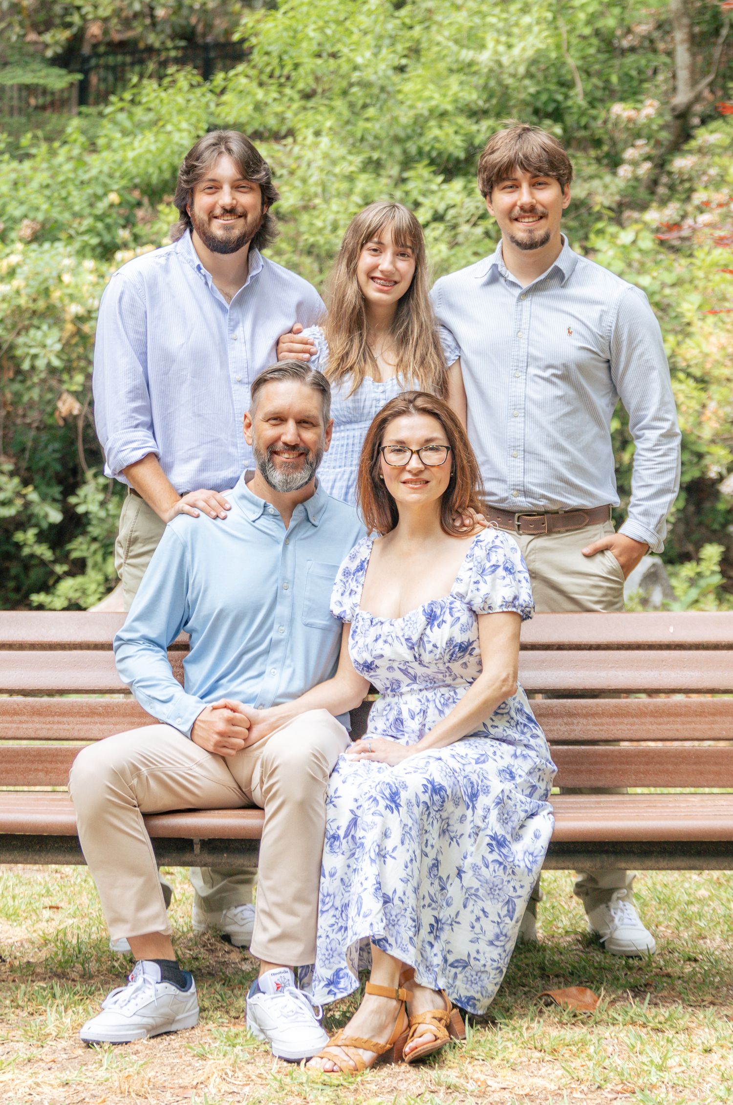Family of five posing on a park bench surrounded by green trees on a sunny day.