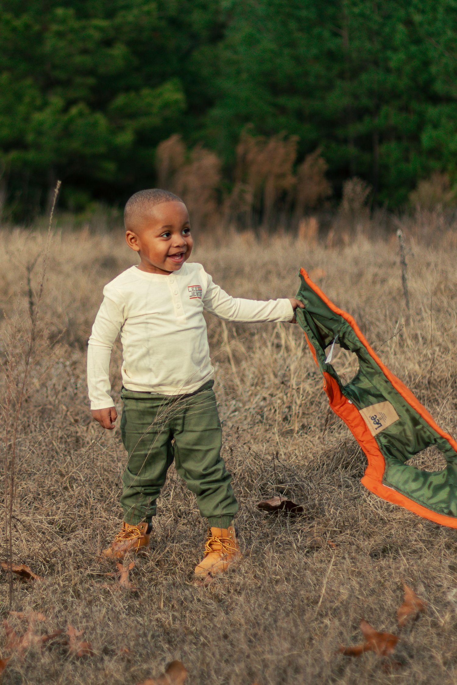 A child in a cream sweater and green pants plays with a bright orange kite in a dry grass field.