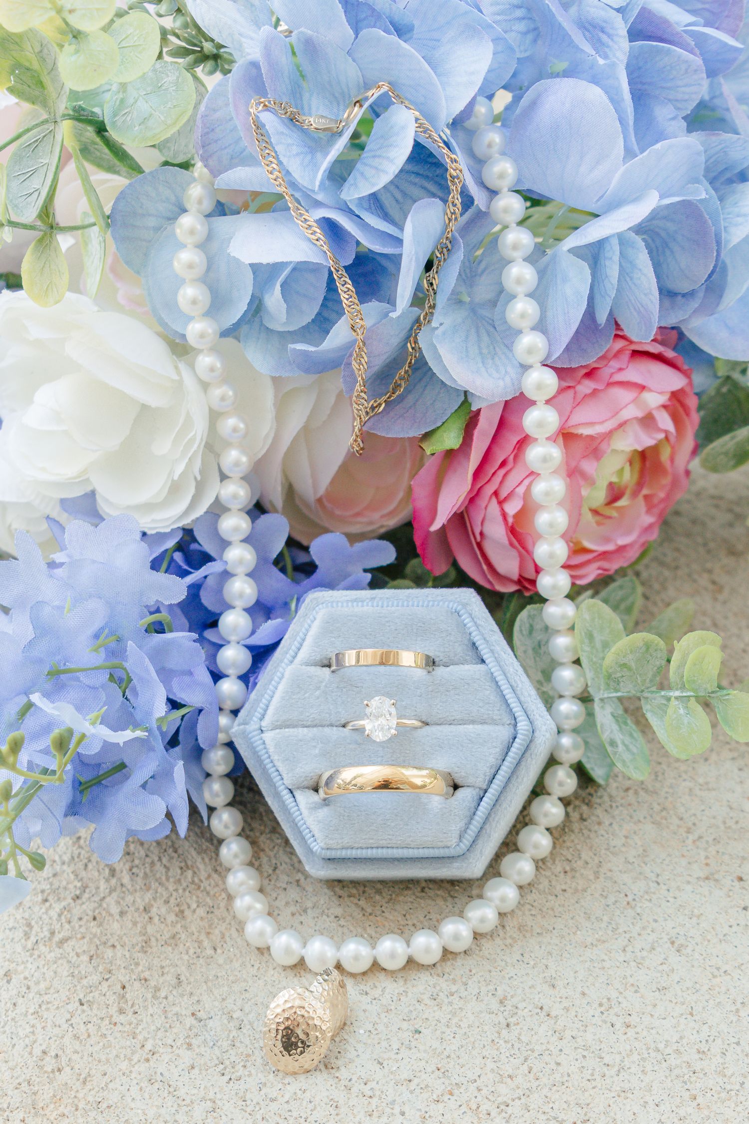 Wedding rings and pearl necklace displayed in a blue velvet box surrounded by colorful flowers.