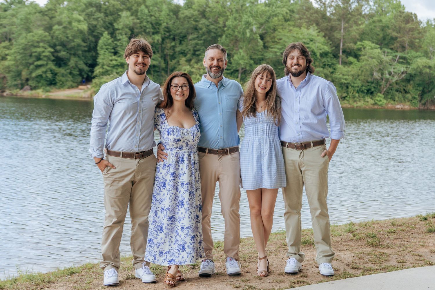 Family of five smiling together on a lakeside shore with lush green trees in the background.