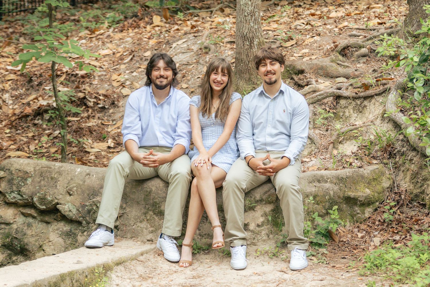 Three young adults sit and smile together on stone steps in a sunlit wooded forest setting.