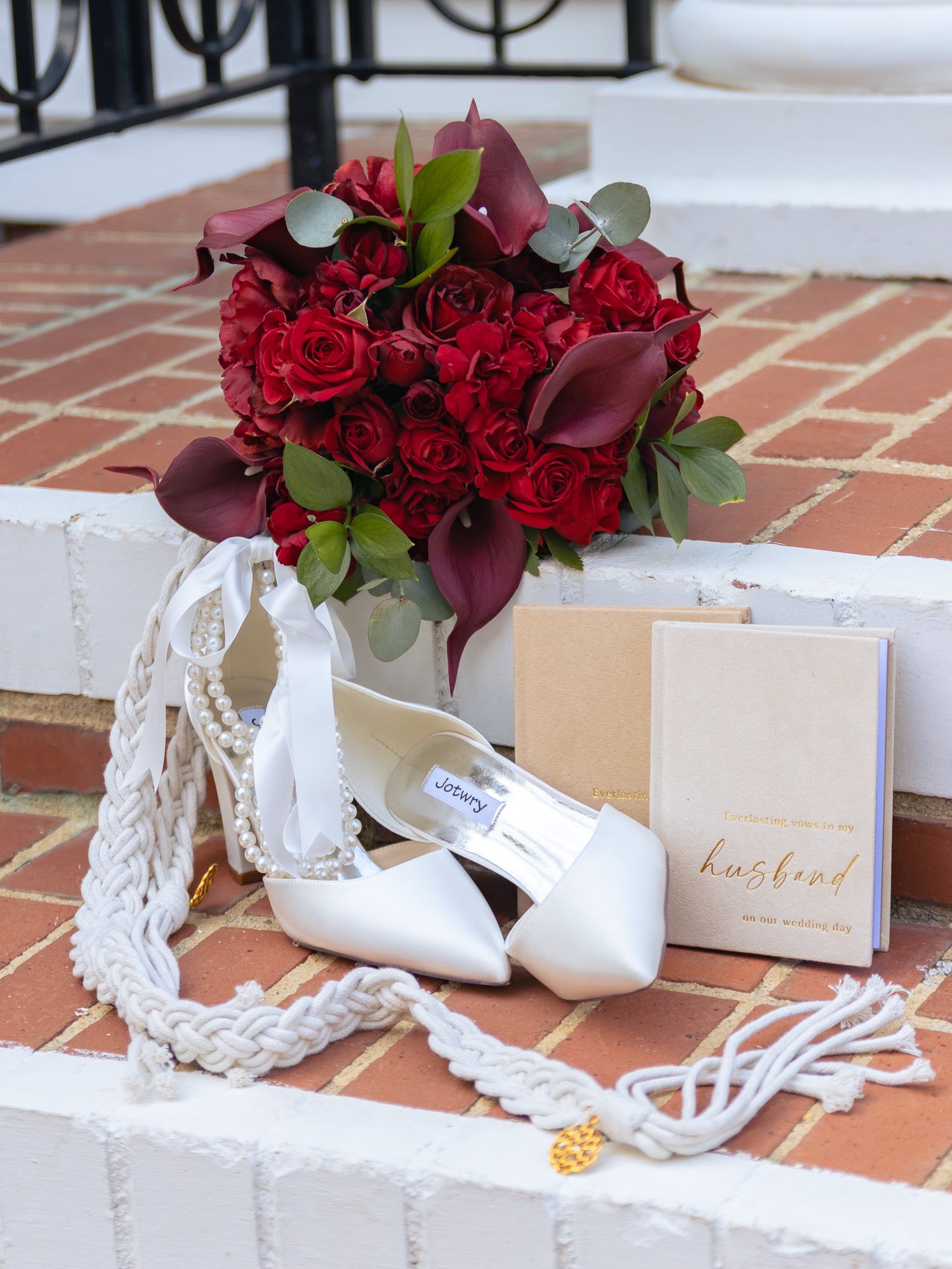 Wedding detail featuring bridal shoes, pearl necklace, handwritten card, and red rose bouquet on brick steps.