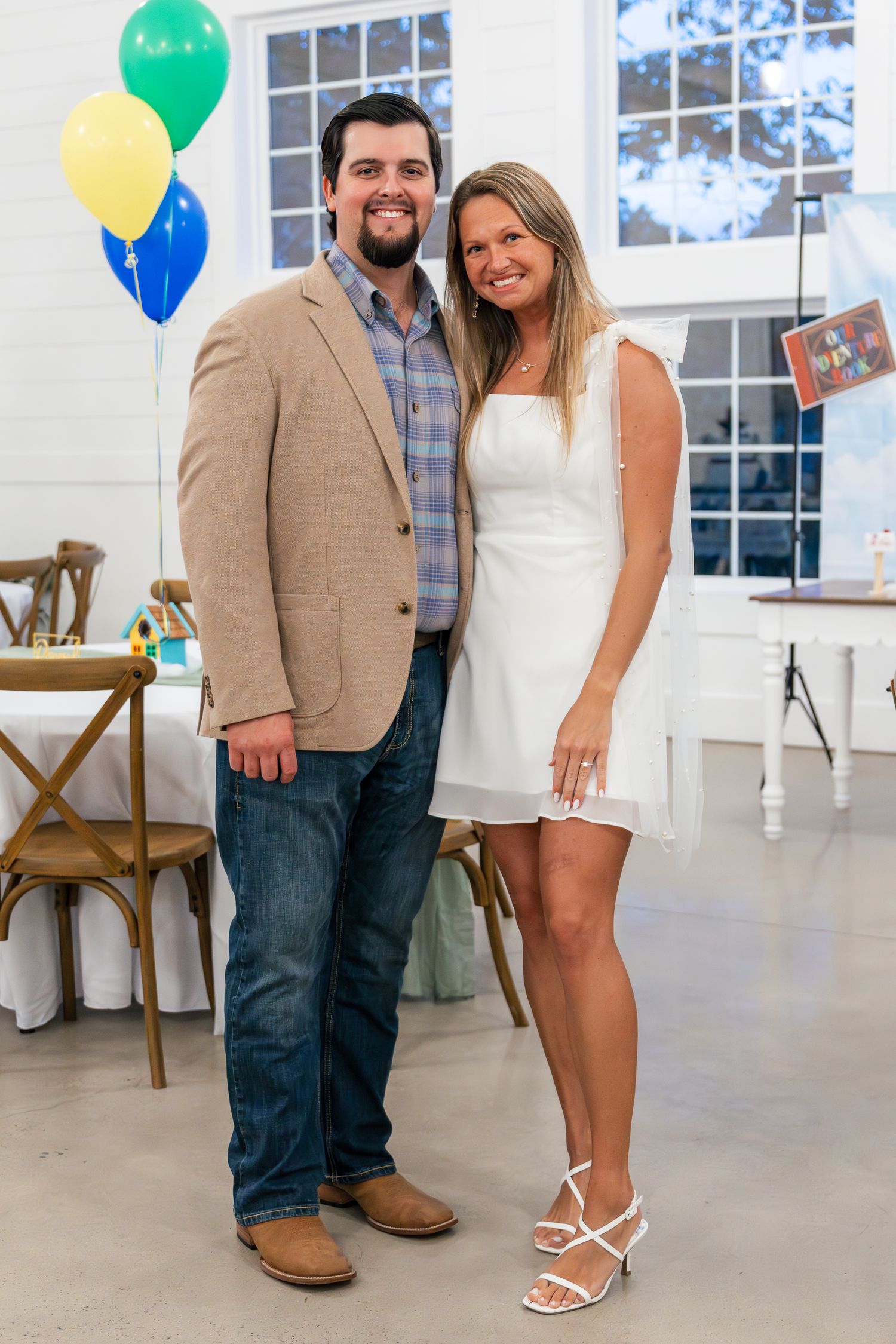 A couple poses together at an indoor event space decorated with colorful balloons and wooden chairs.