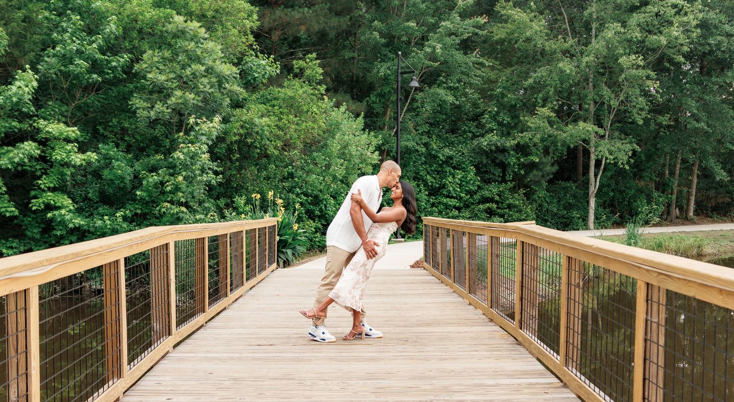 Romantic couple sharing a dip kiss on a wooden bridge surrounded by lush greenery.