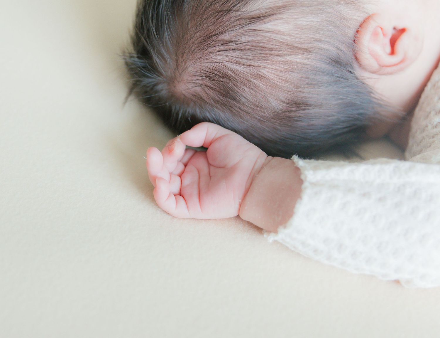 Close-up of a sleeping newborn baby's tiny hand and dark hair on a soft cream background.