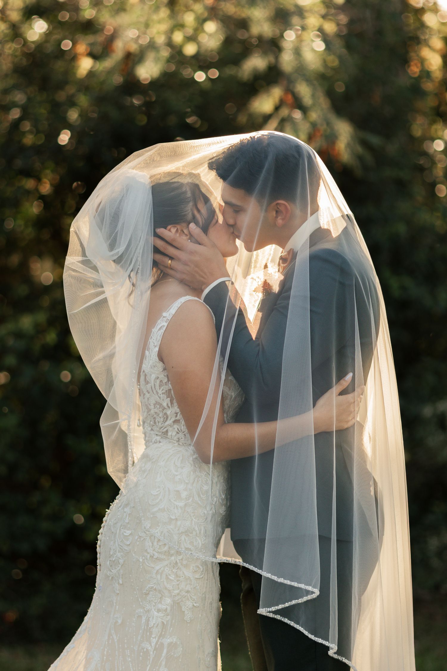 Bride and groom sharing a kiss under a flowing white veil outdoors in golden sunlight.