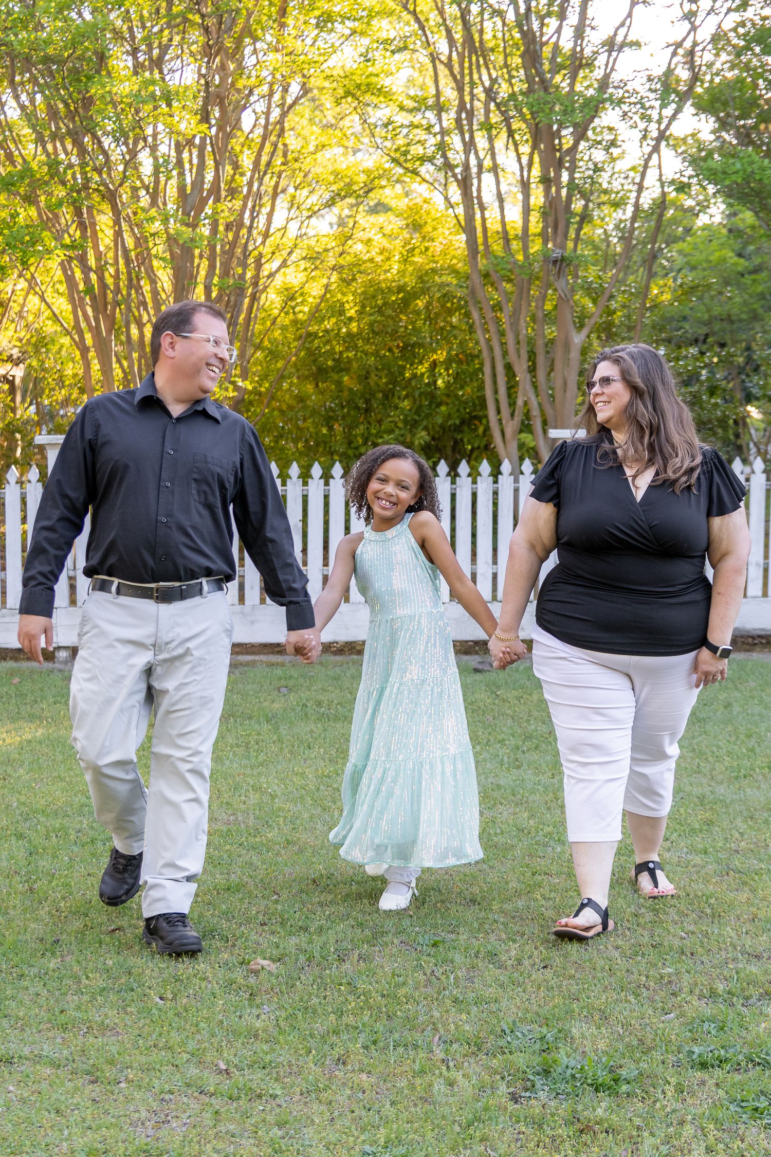 A happy family of three holds hands while walking across a green lawn in warm golden evening light.
