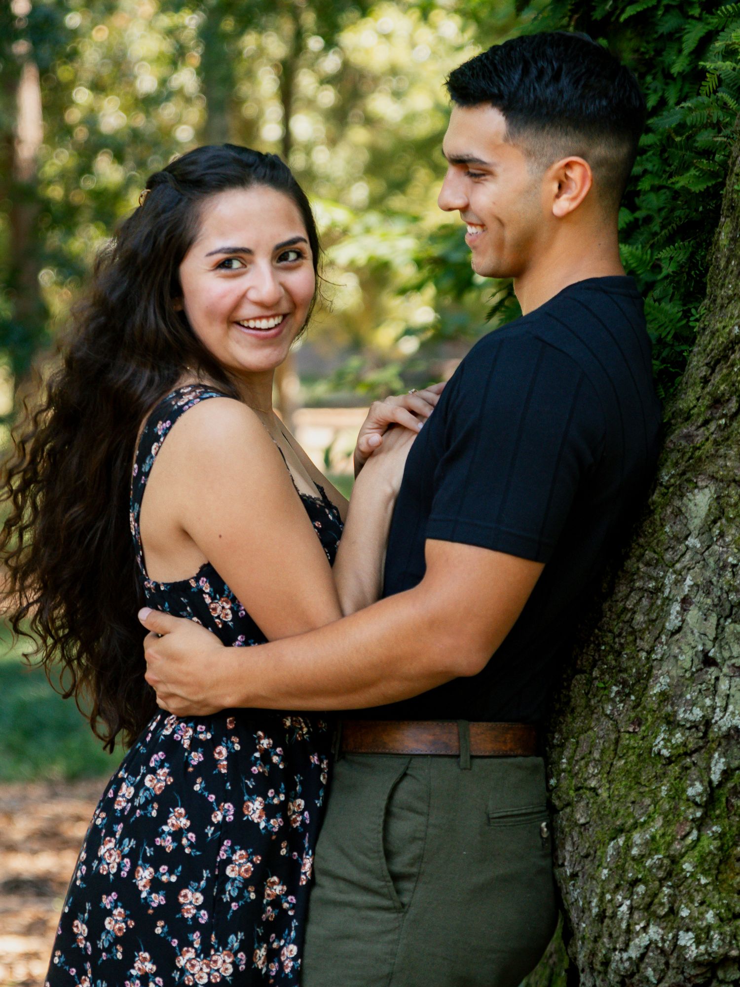 Smiling couple embracing near a tree in a sunlit forest during an outdoor engagement session.