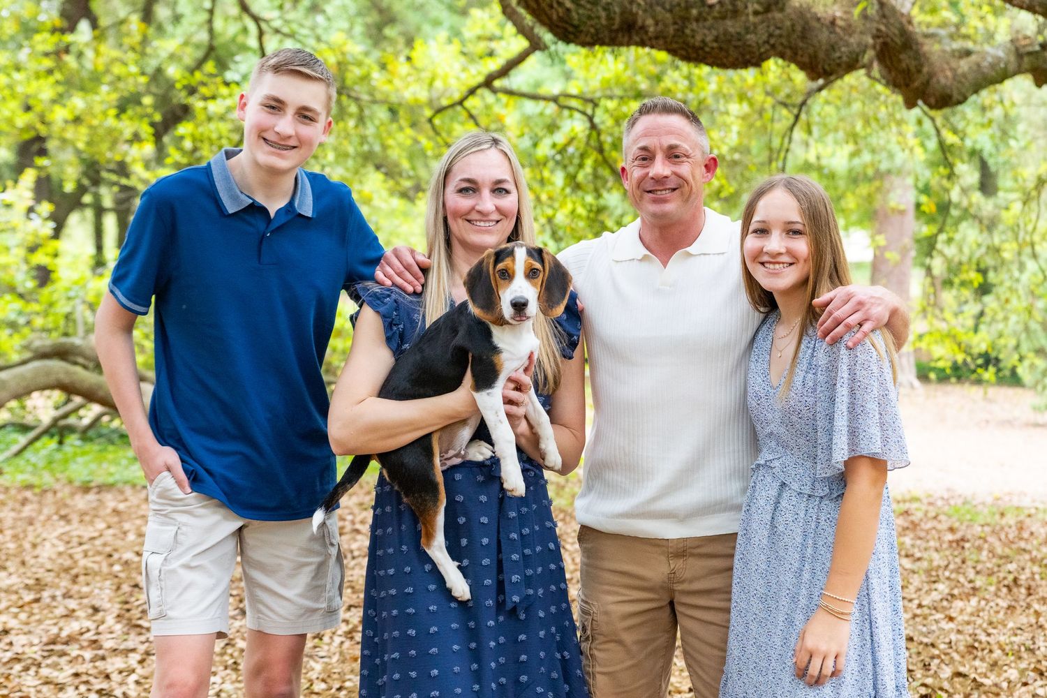 A smiling family of four poses outdoors under a large tree while holding an adorable beagle puppy.