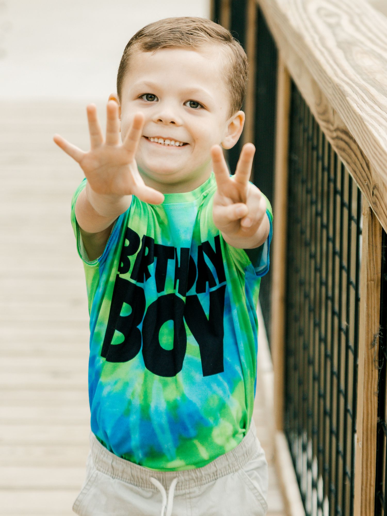 A cheerful young boy in a tie-dye Birthday Boy shirt holds up seven fingers on a wooden staircase.