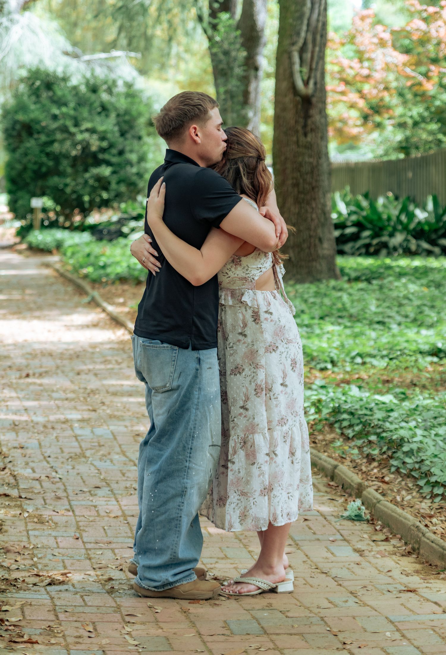 Couple sharing a romantic kiss while embracing on a brick garden path surrounded by lush green trees.