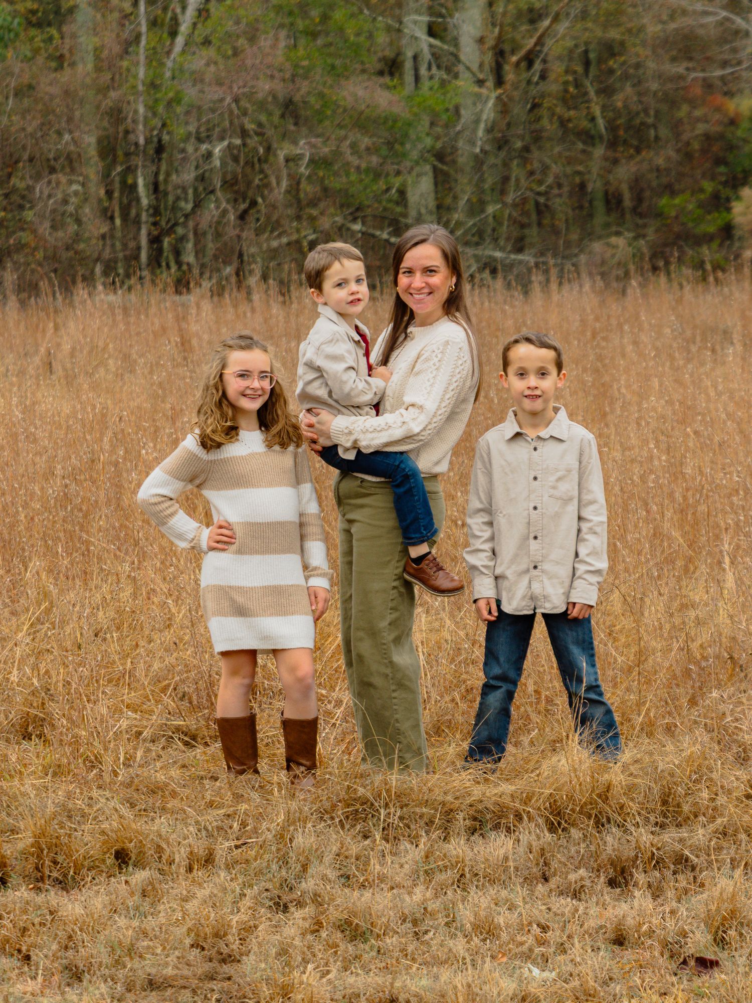 A smiling mother holds a toddler while standing with two older children in a golden autumn field.