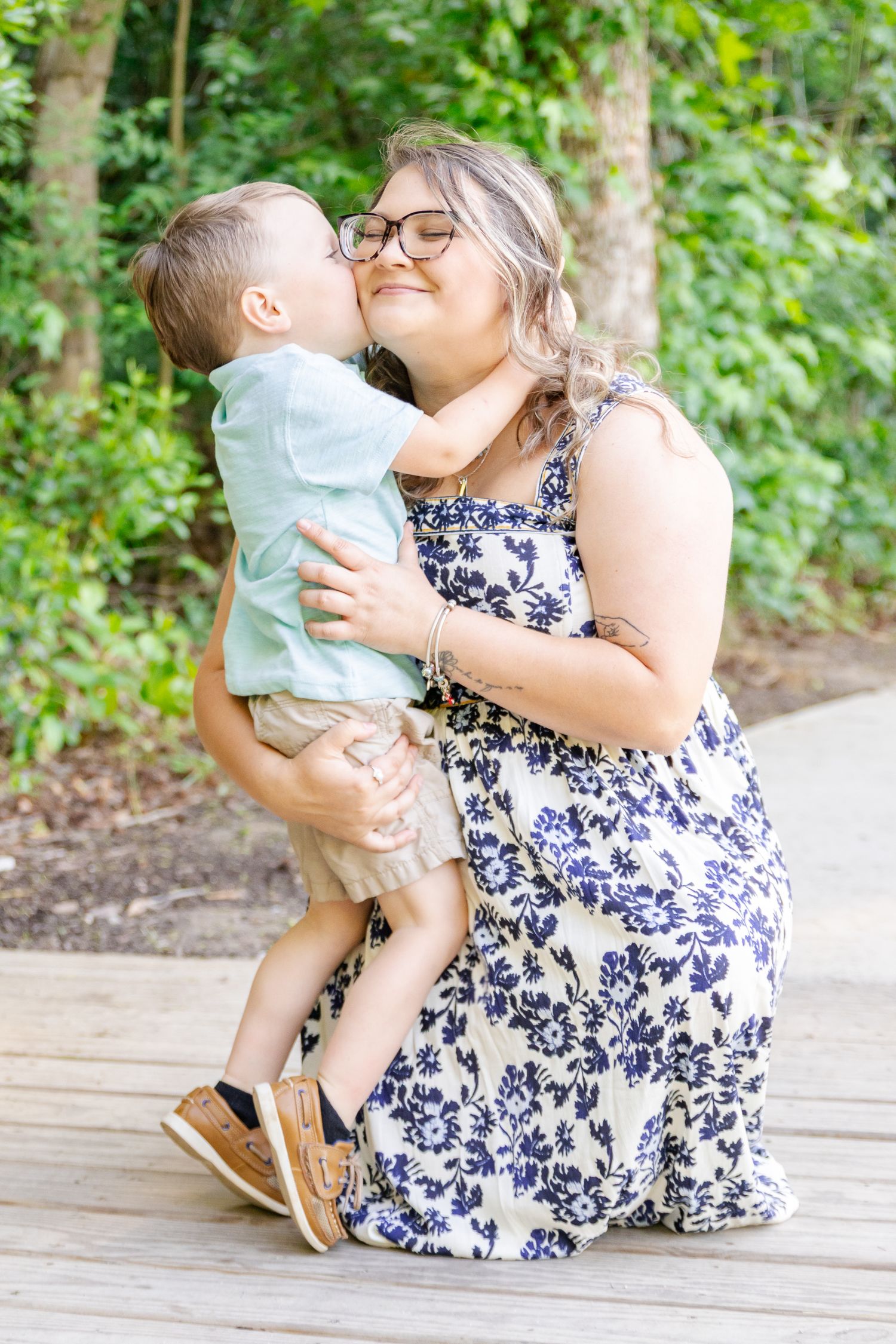 A mother kneeling on a wooden deck holds her young son as he gives her a sweet kiss on the cheek outdoors.