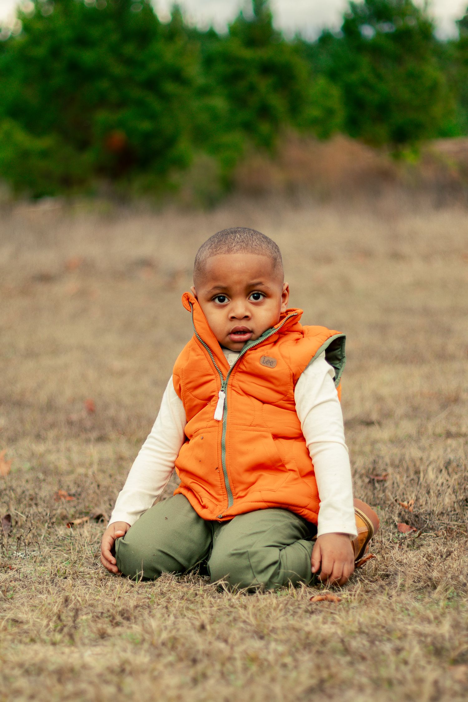 A young child sits in a field wearing an orange puffy vest and cream colored long sleeve shirt.