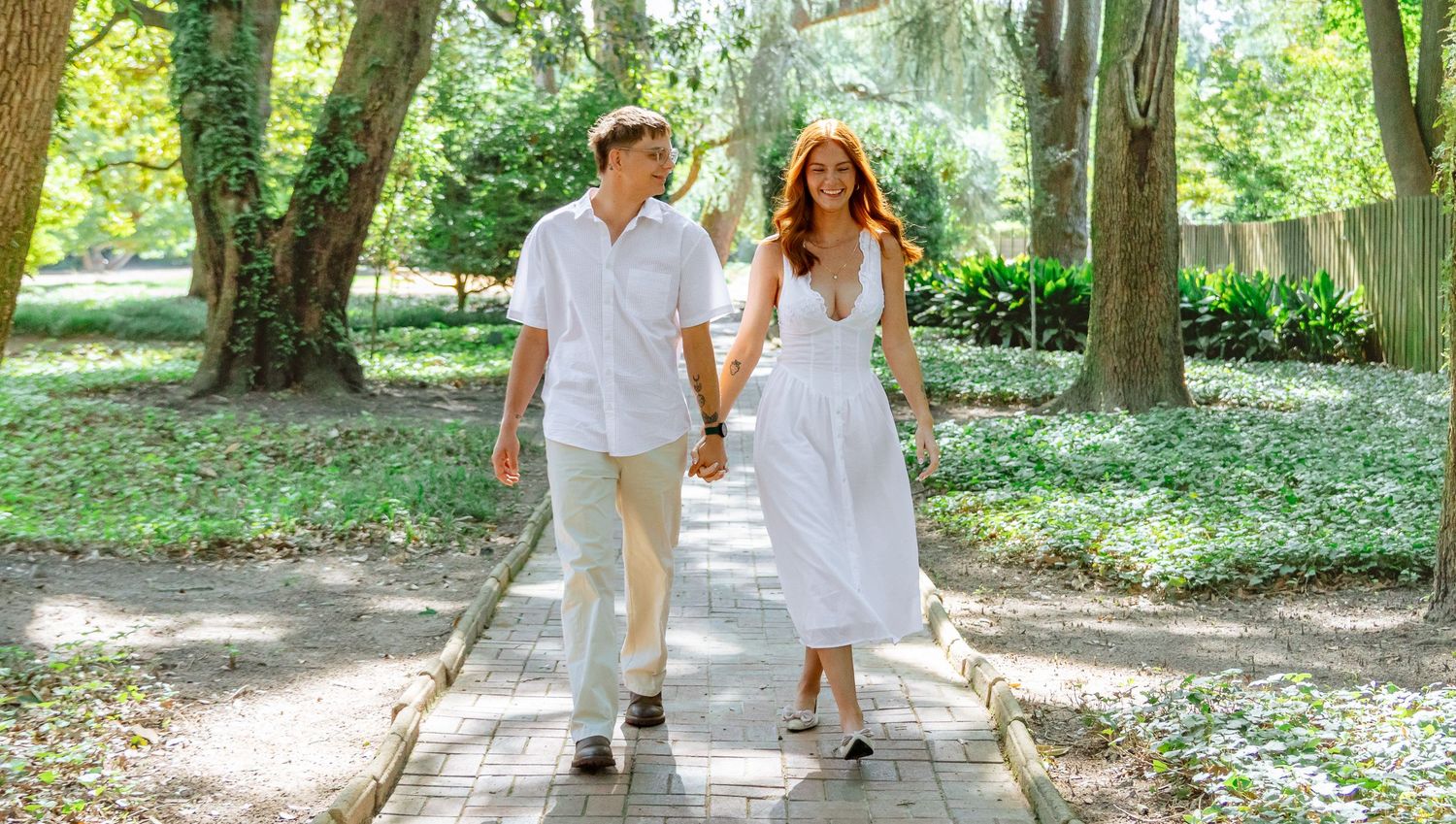 Engaged couple walking hand in hand along a tree-lined Hopeland Gardens path in Aiken, South Carolina.
