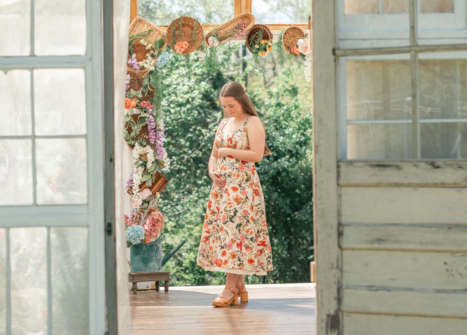 Pregnant woman in floral dress standing in rustic doorway surrounded by greenery and decorative hats.