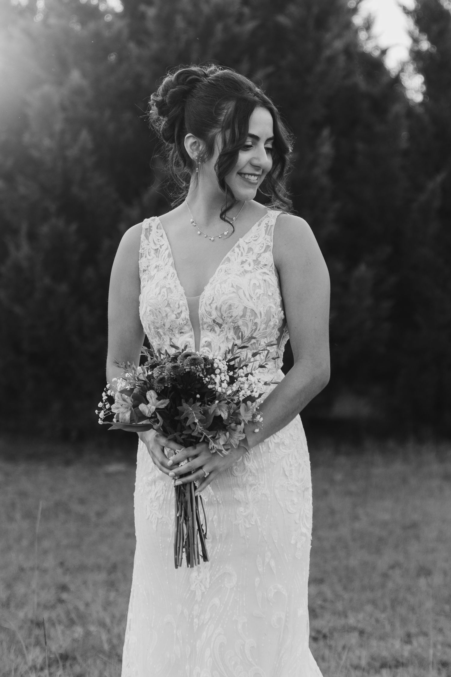 A bride in a lace V-neck wedding dress smiles while holding a rustic bouquet in black and white photography.