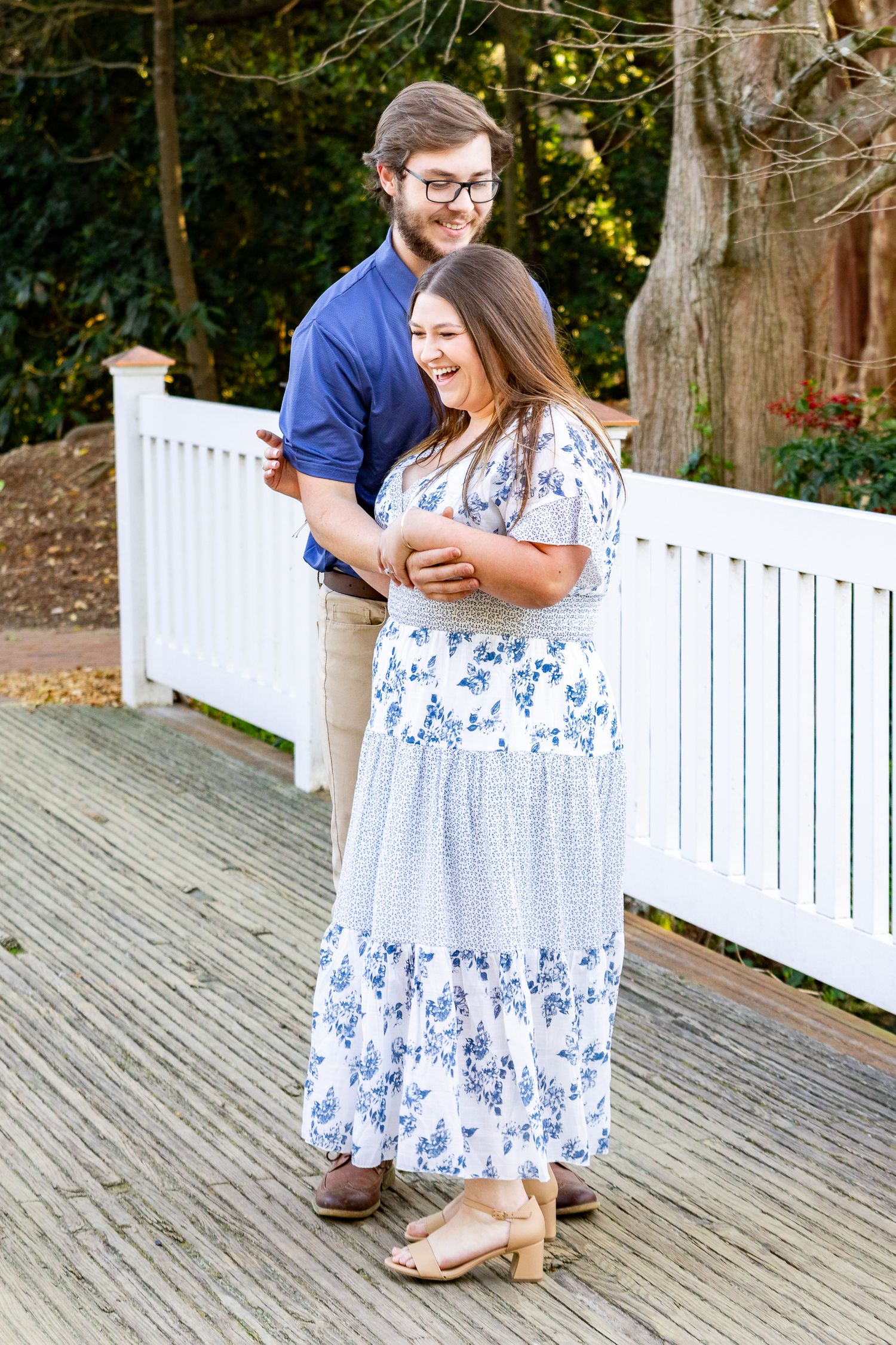 Happy couple standing on a wooden deck with a white fence, woman laughing in floral dress.