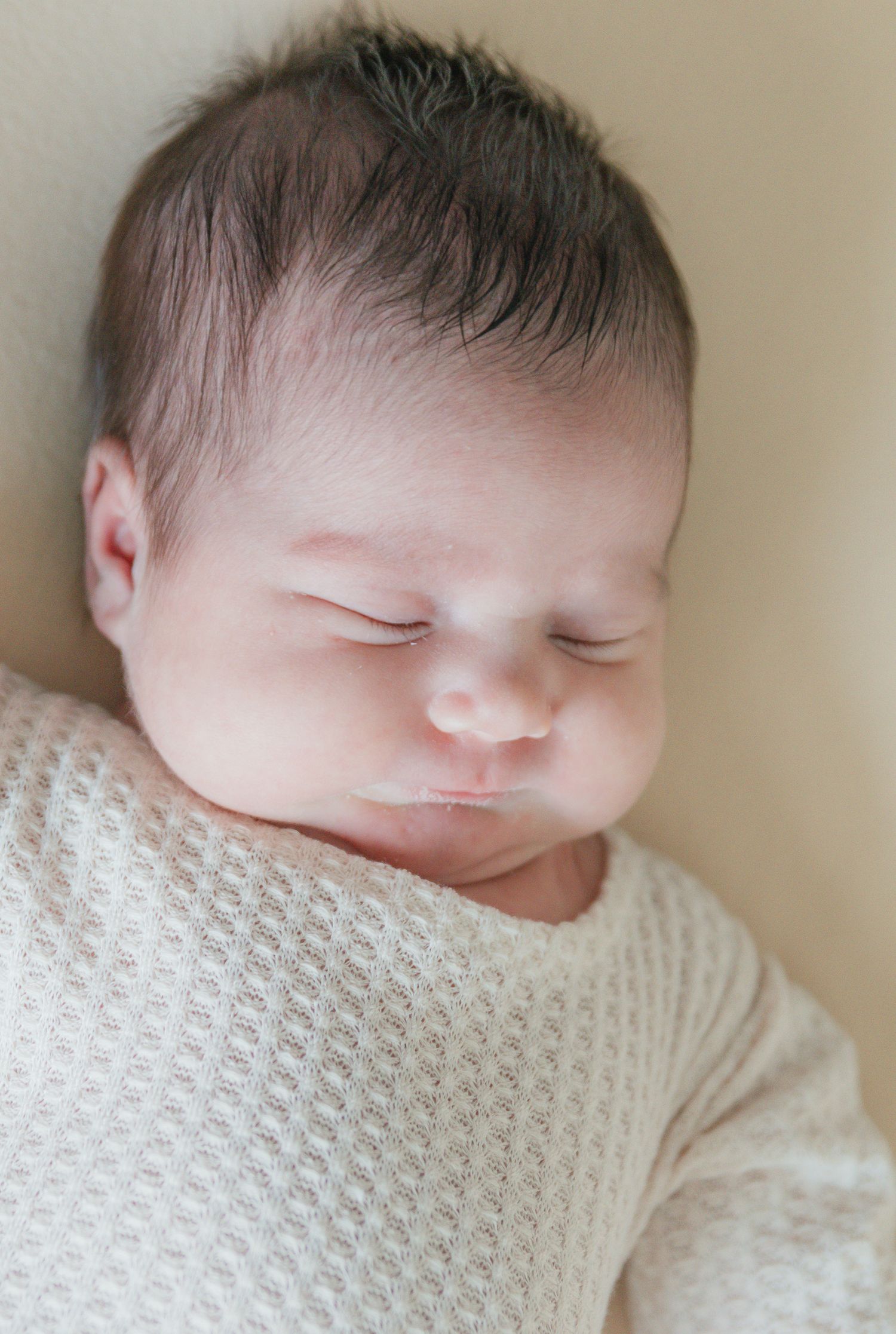 Close-up portrait of a peaceful sleeping newborn baby with dark hair in a soft white knit outfit.