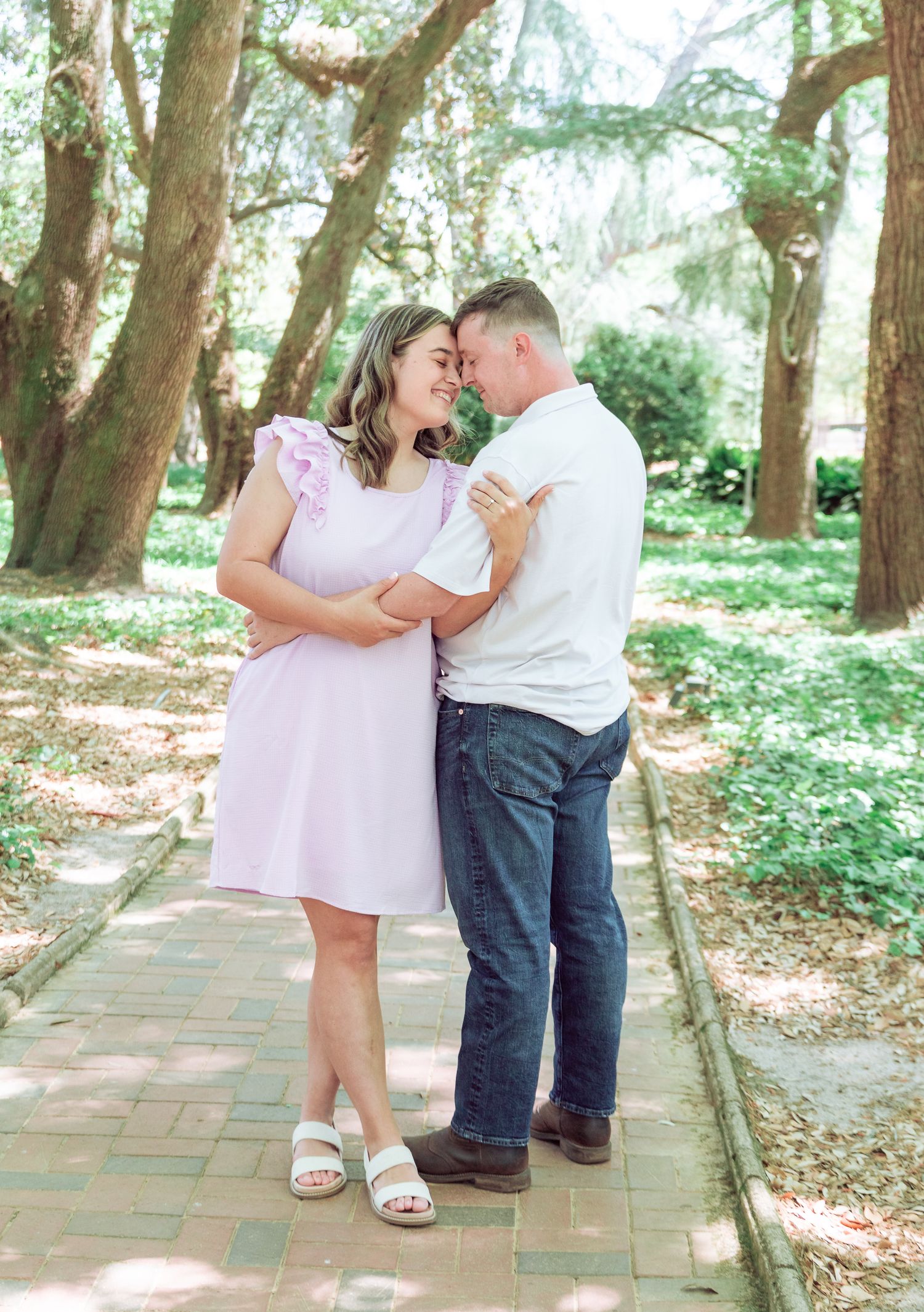 Couple sharing a romantic moment on a tree-lined brick path, woman in pink dress and man in white shirt.