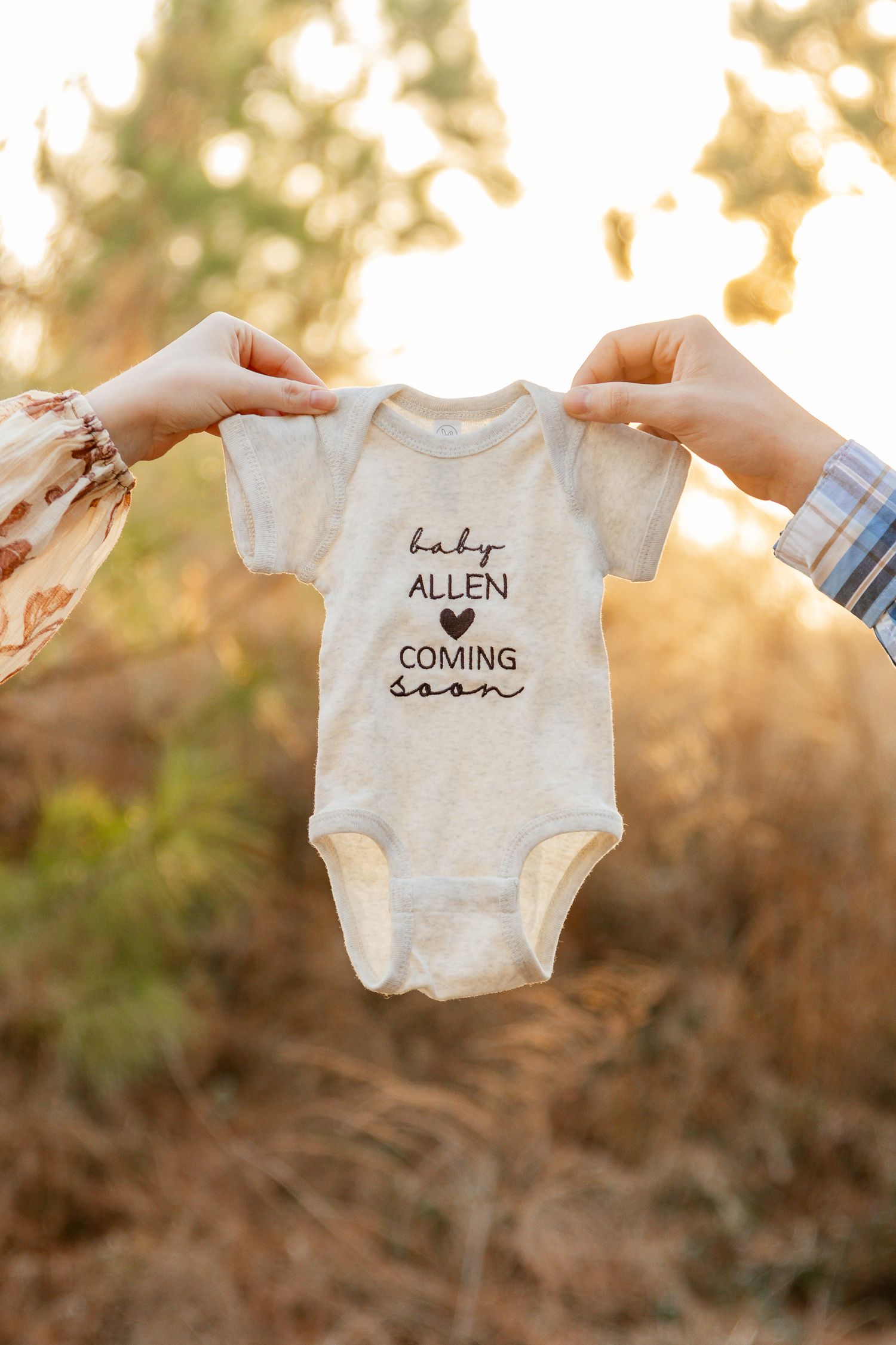 Two hands hold up a white baby onesie with text 'baby Allen coming soon' against a golden sunset background.