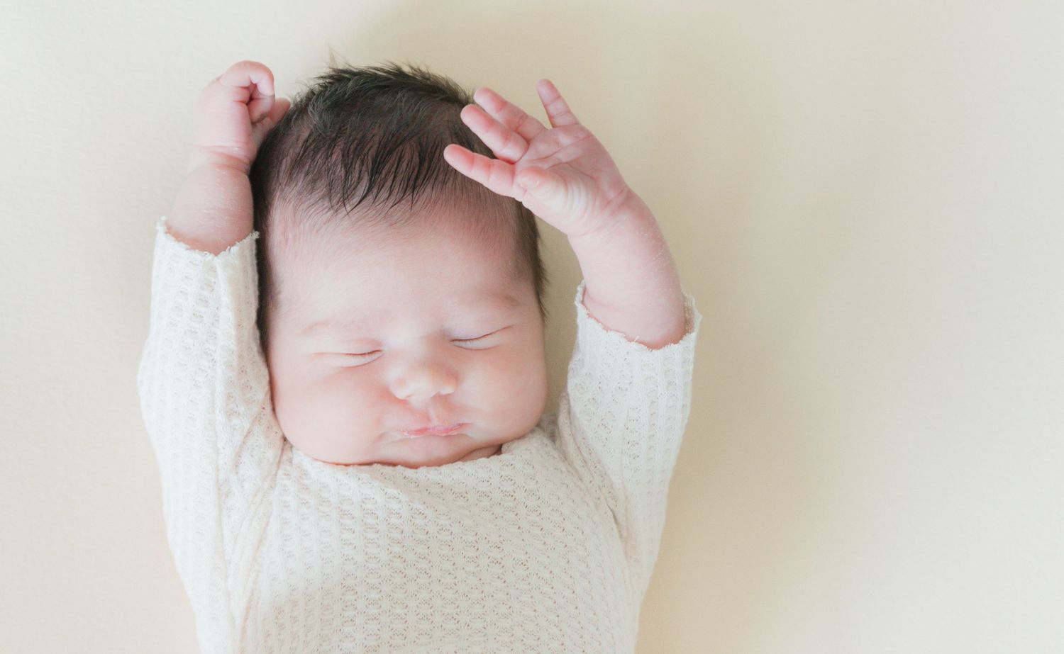 Adorable newborn baby sleeping in white knit outfit with tiny hands raised above head.