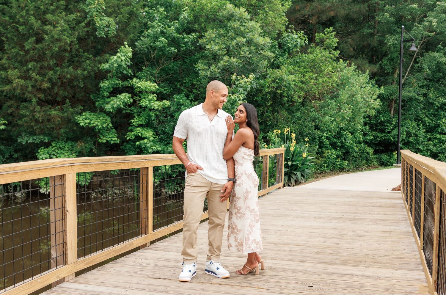 Couple standing together on a wooden bridge surrounded by lush green trees, woman in floral dress, man in white shirt.