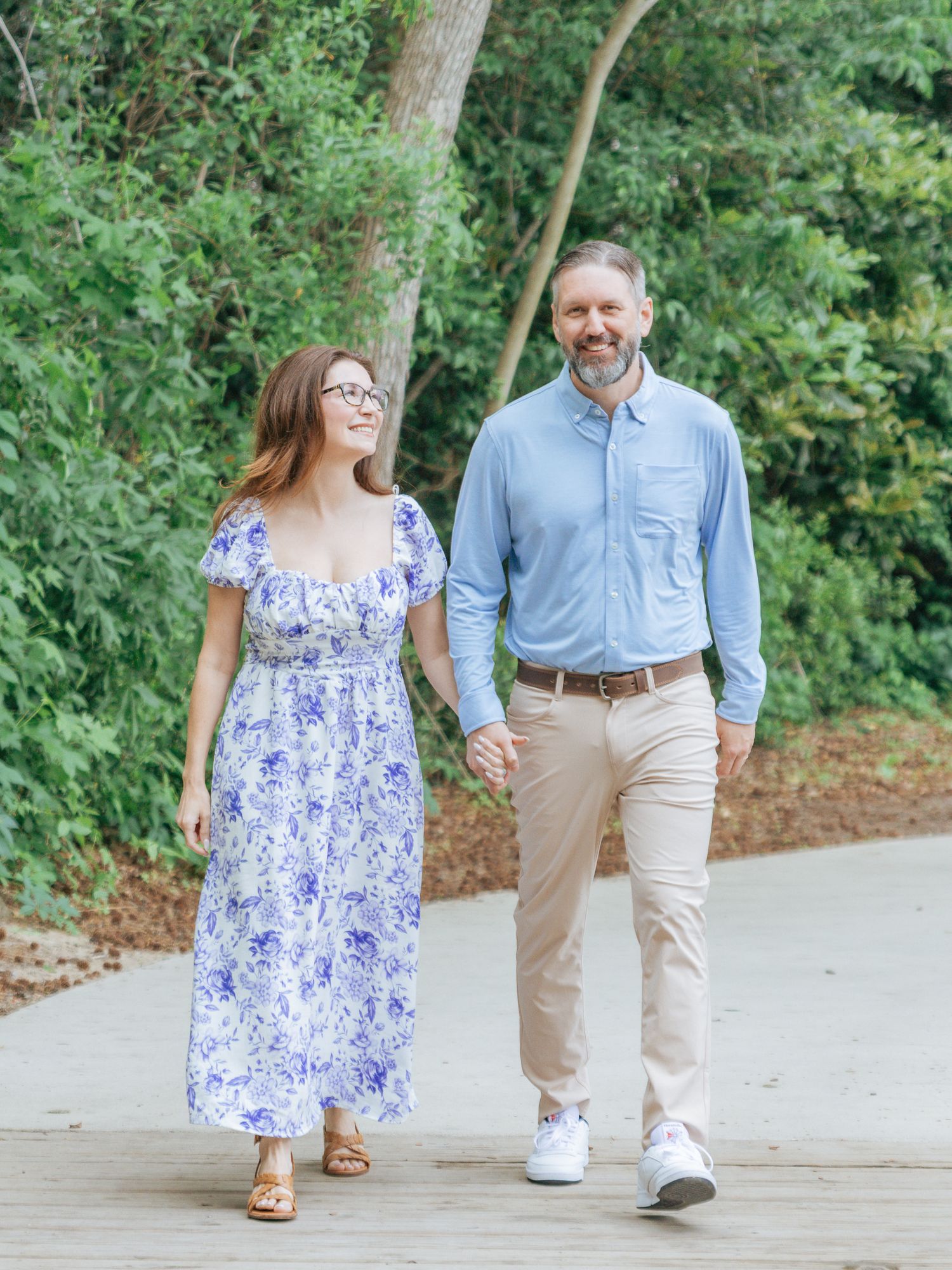 Smiling couple walking hand in hand on a wooden path surrounded by lush green trees.