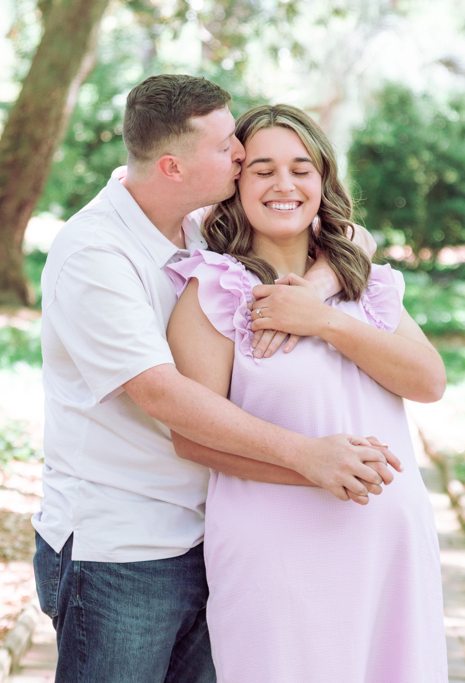 Smiling pregnant woman in lavender dress embraced by her partner outdoors.