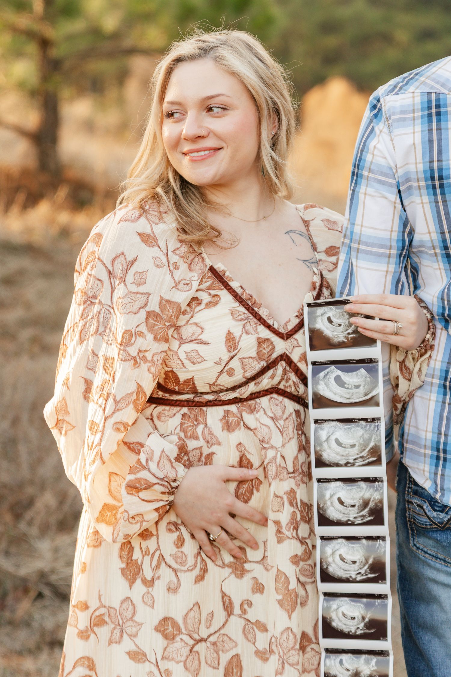 Expectant person in floral dress holds ultrasound photos while standing outdoors during golden hour.
