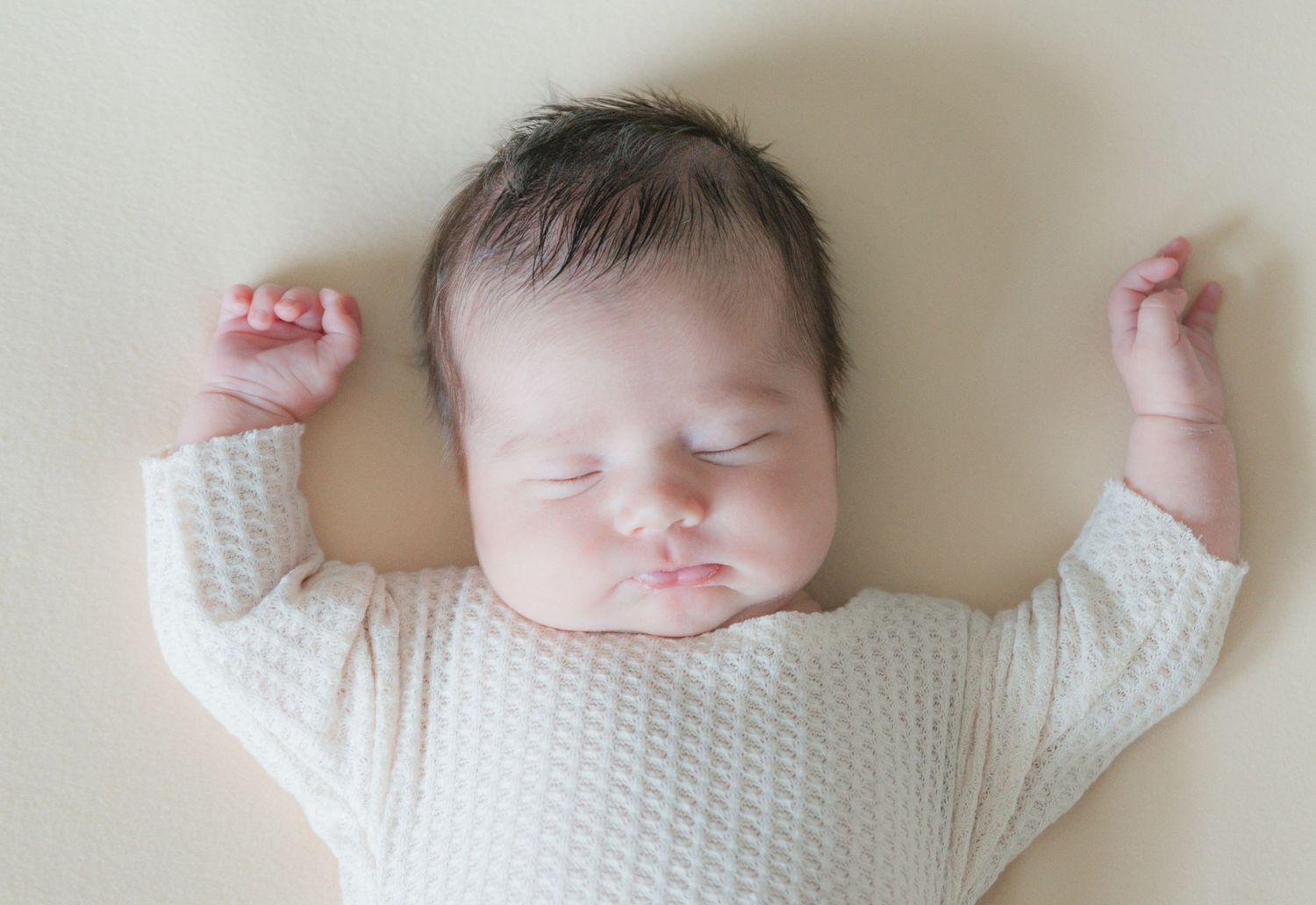 Peaceful newborn baby sleeping with arms raised wearing a white knit onesie on a cream background.