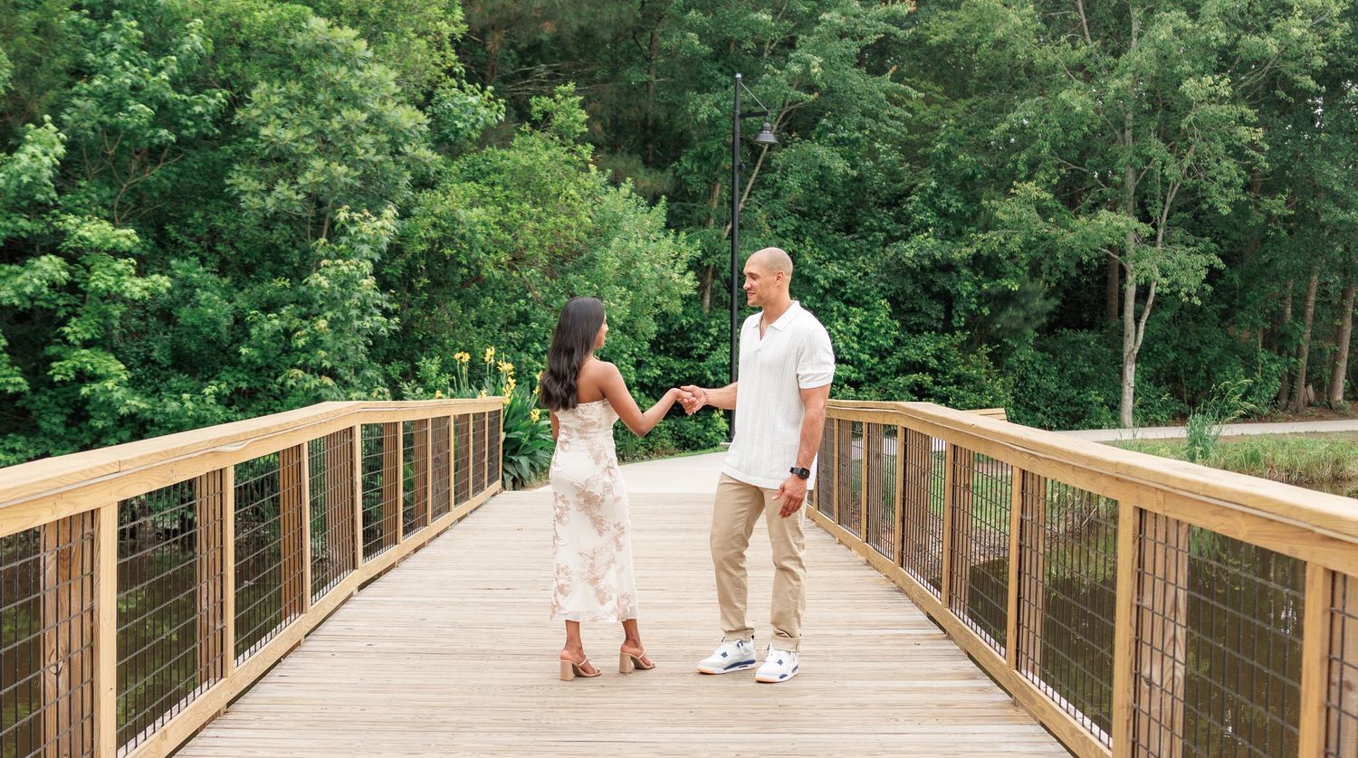 Couple holding hands facing each other on a wooden footbridge surrounded by greenery.