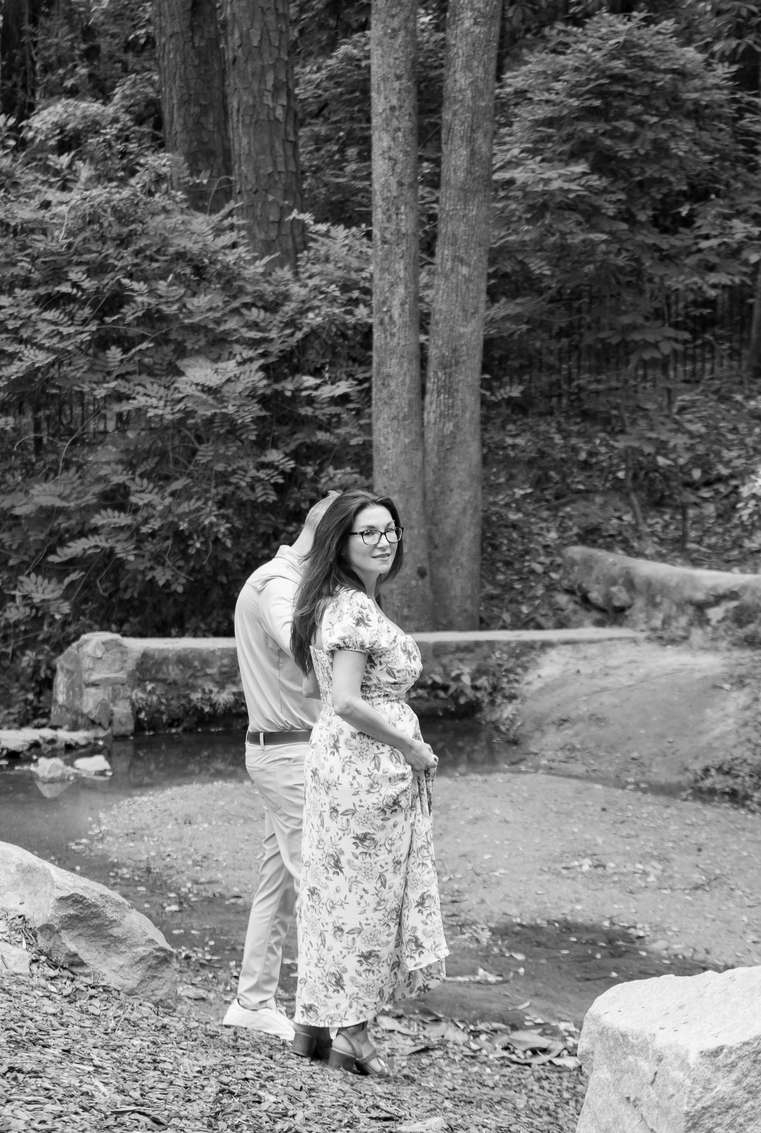 Black and white photo of a couple embracing near a creek surrounded by trees and rocks.
